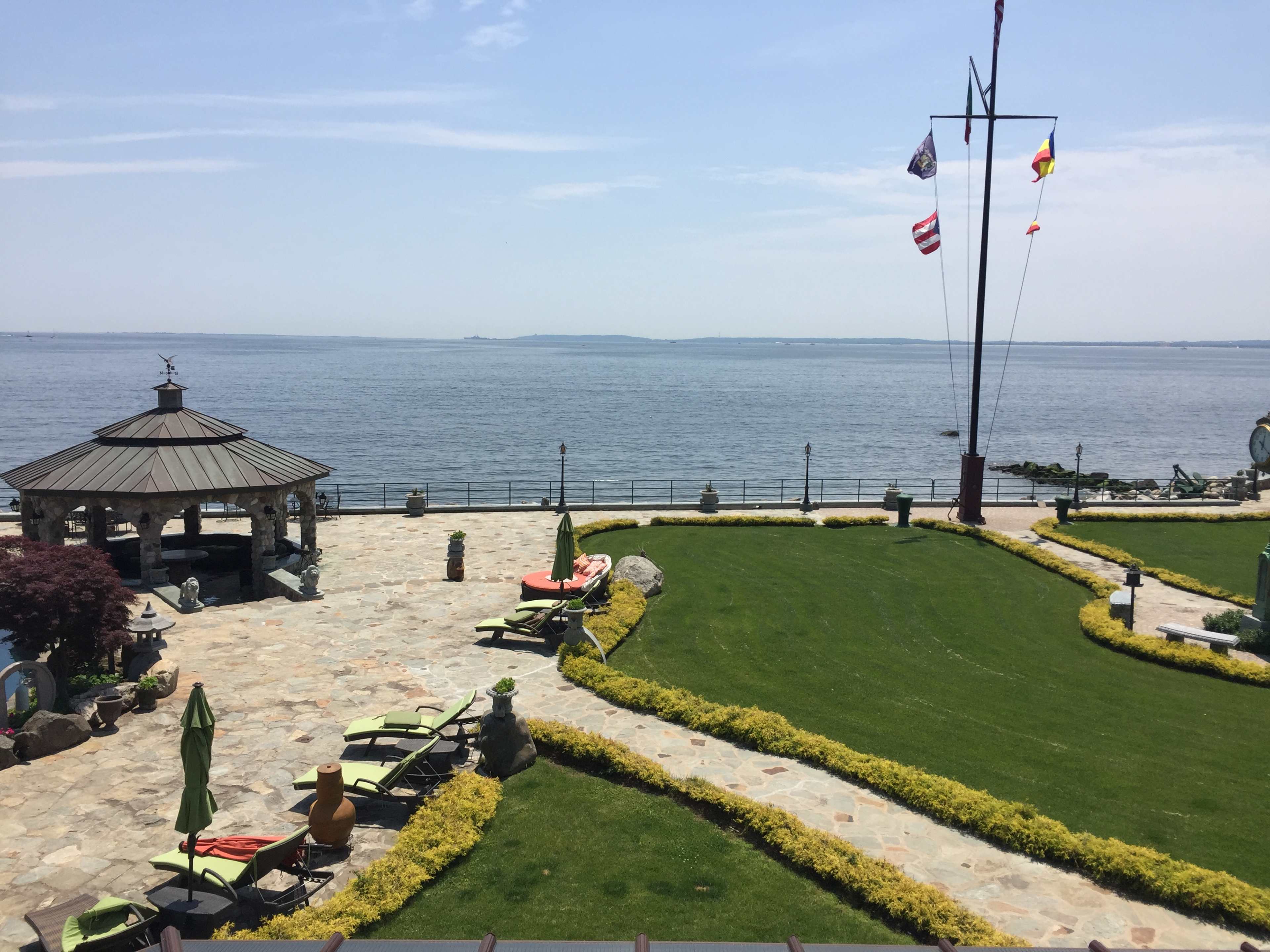 The image shows a stone-paved terrace overlooking a body of water, with lounge chairs and flags nearby.