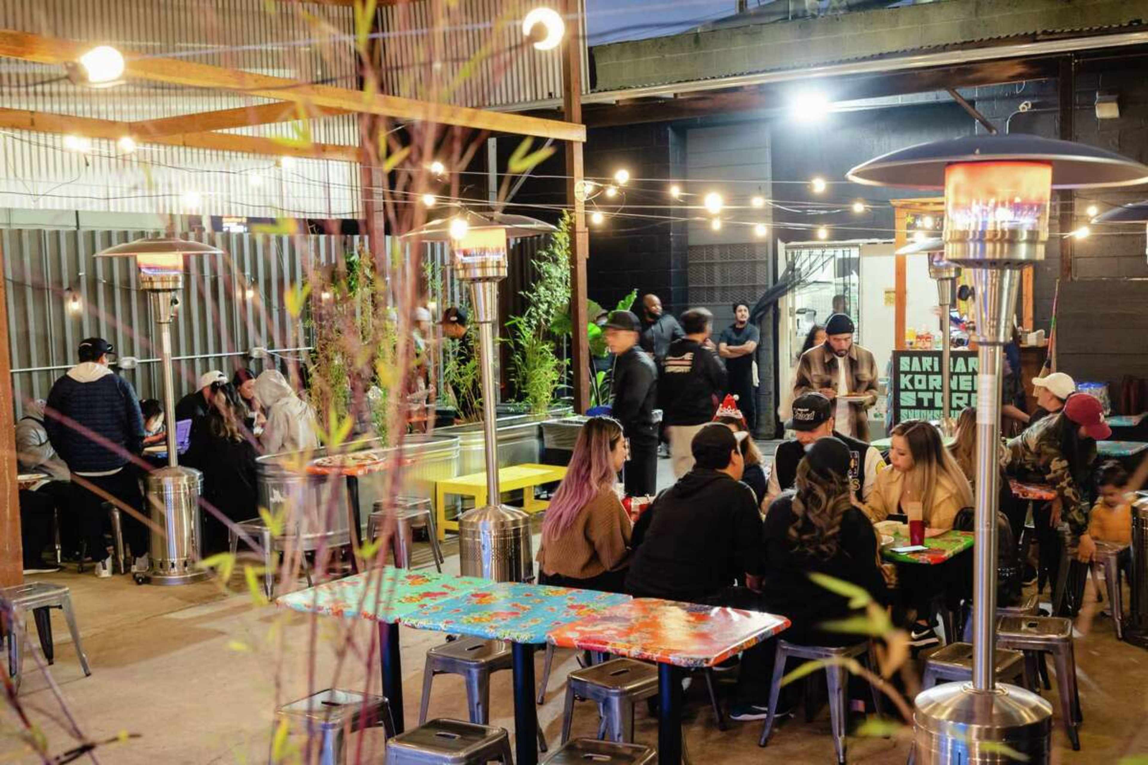 A crowded outdoor dining area features colorful tables, string lights, and heating lamps, with patrons seated and socializing.