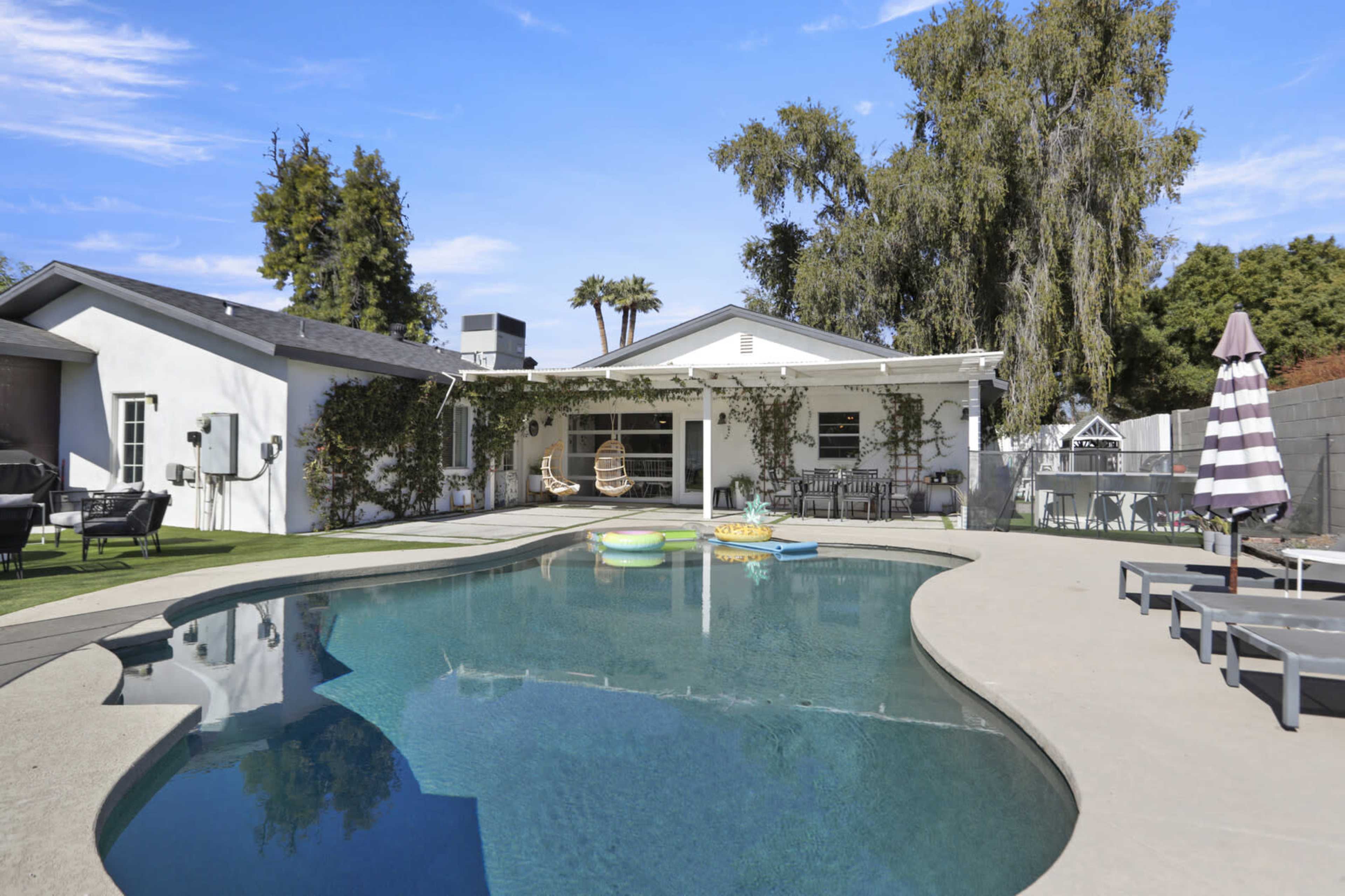 A backyard features a pool with floating objects, surrounded by lounging chairs and a covered patio with outdoor seating.