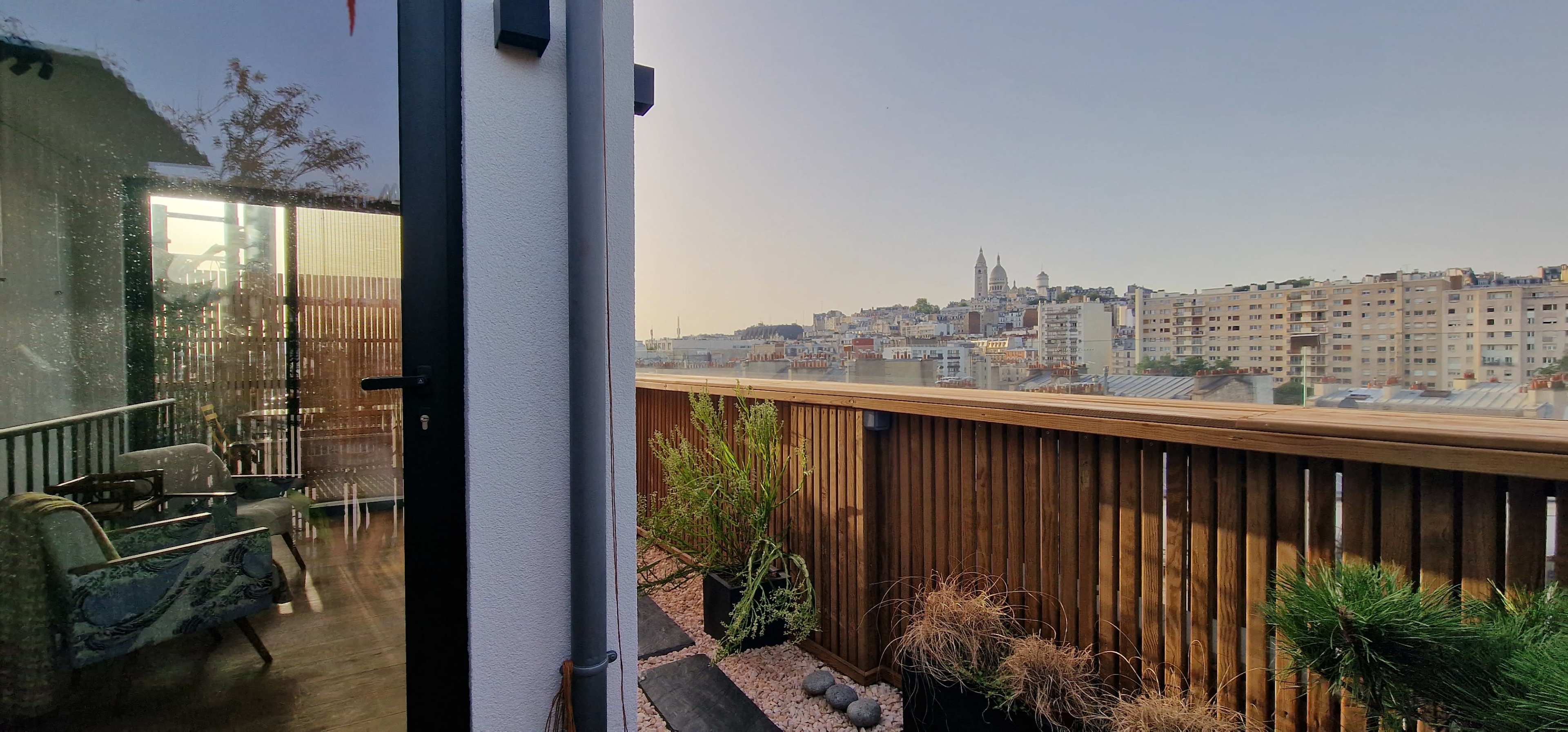 A view from a terrace shows a cityscape with buildings and a distant church spire under a clear sky.