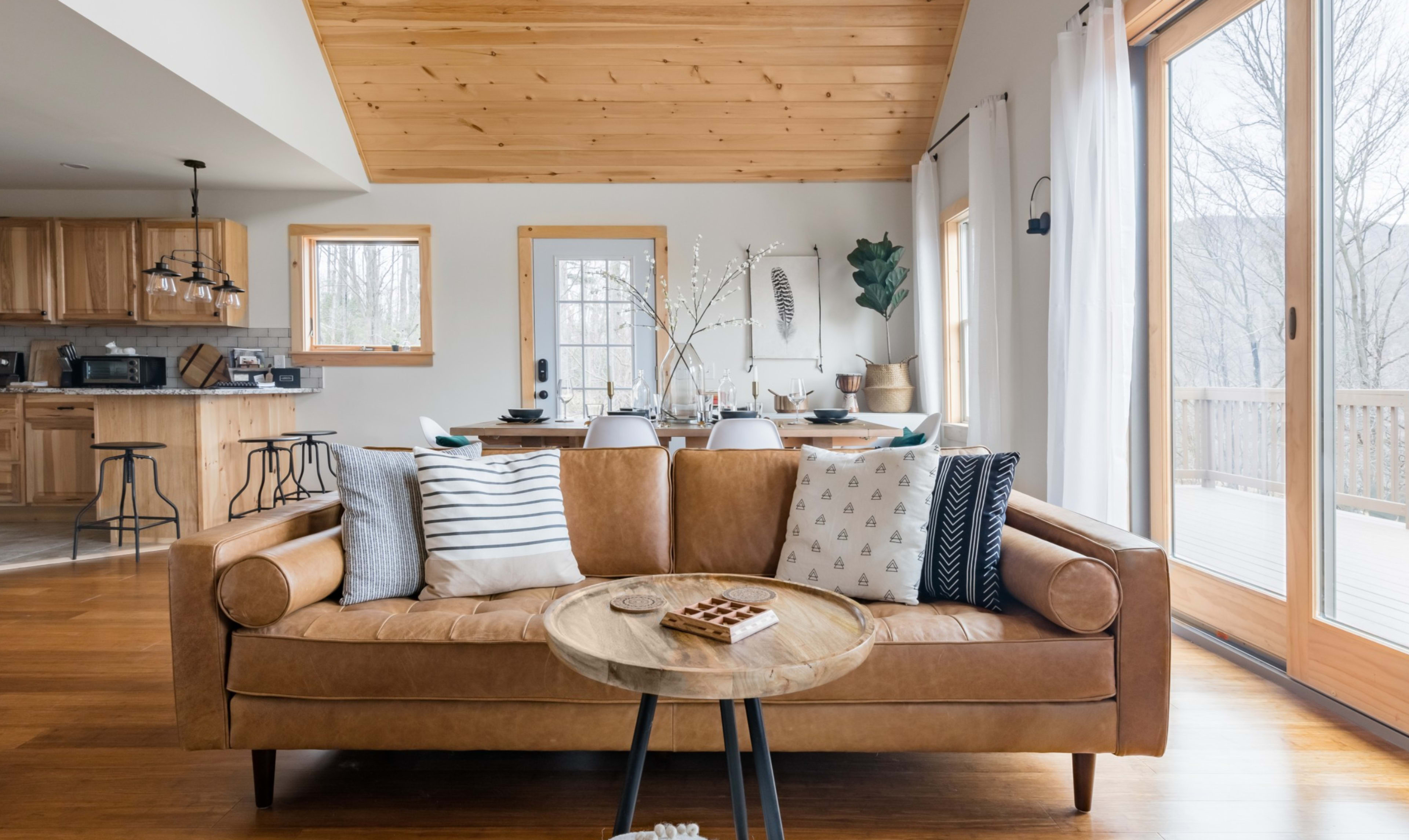 A cozy living room with a leather sofa, a wooden coffee table, and large windows leading to a deck, with a kitchen visible in the background.