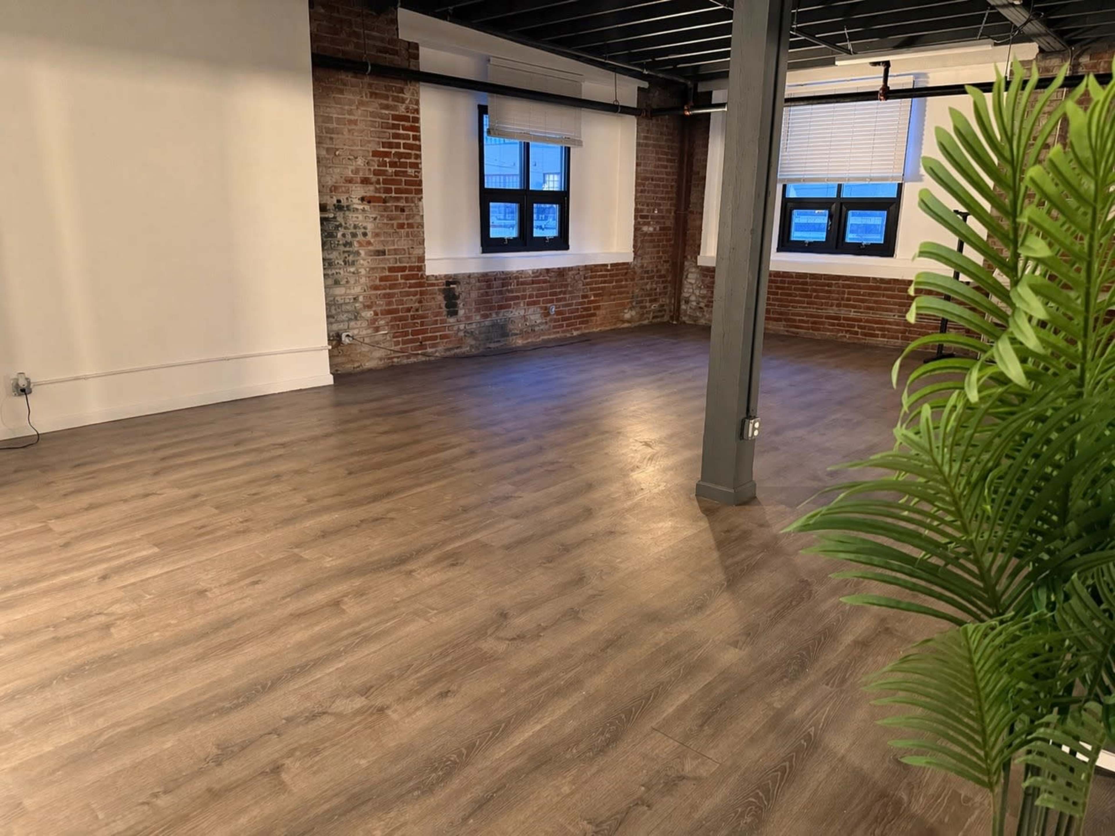 The image shows an empty room with wooden flooring, exposed brick walls, and large windows.
