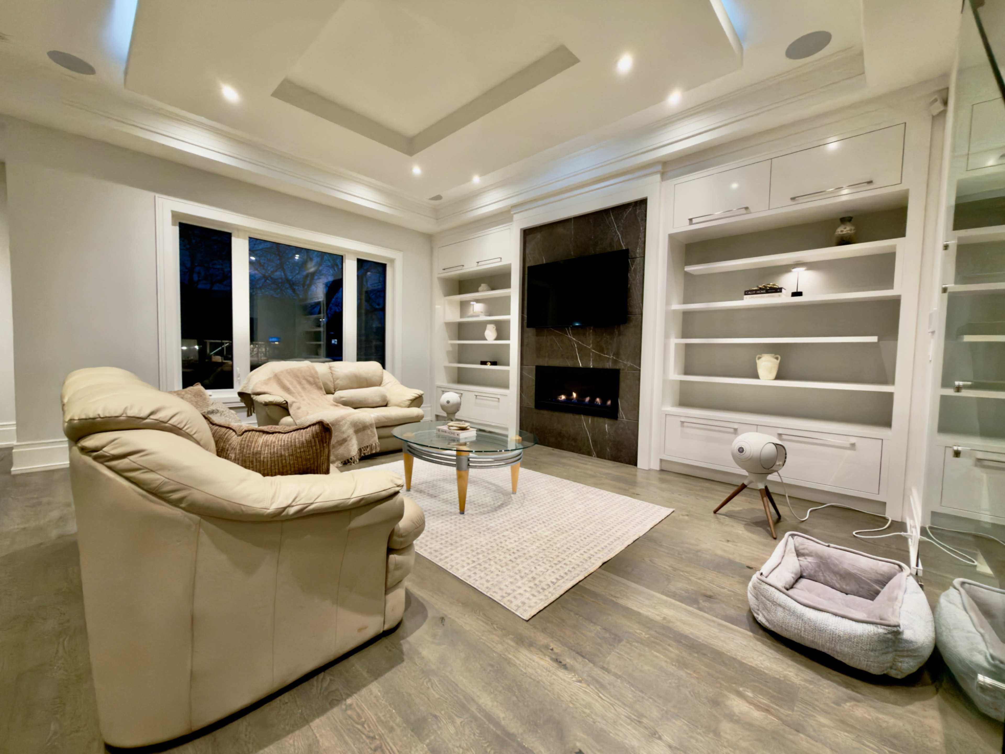 The living room features a set of beige armchairs, a glass coffee table, a modern fireplace, and built-in shelves against light-colored walls.