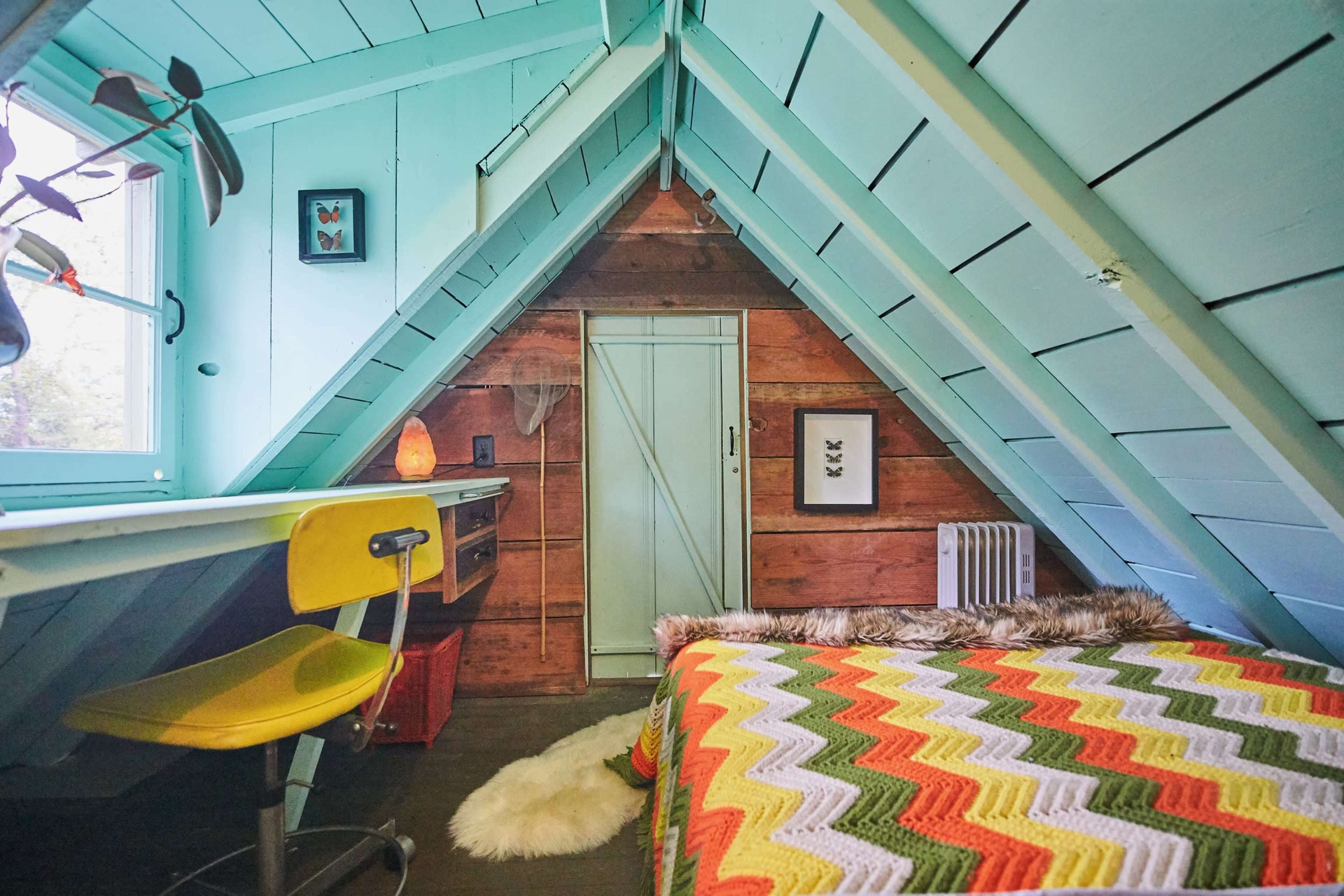 A cozy attic room with a sloped ceiling, featuring a patterned bedspread, a small desk with a yellow chair, and wooden accents on the walls.