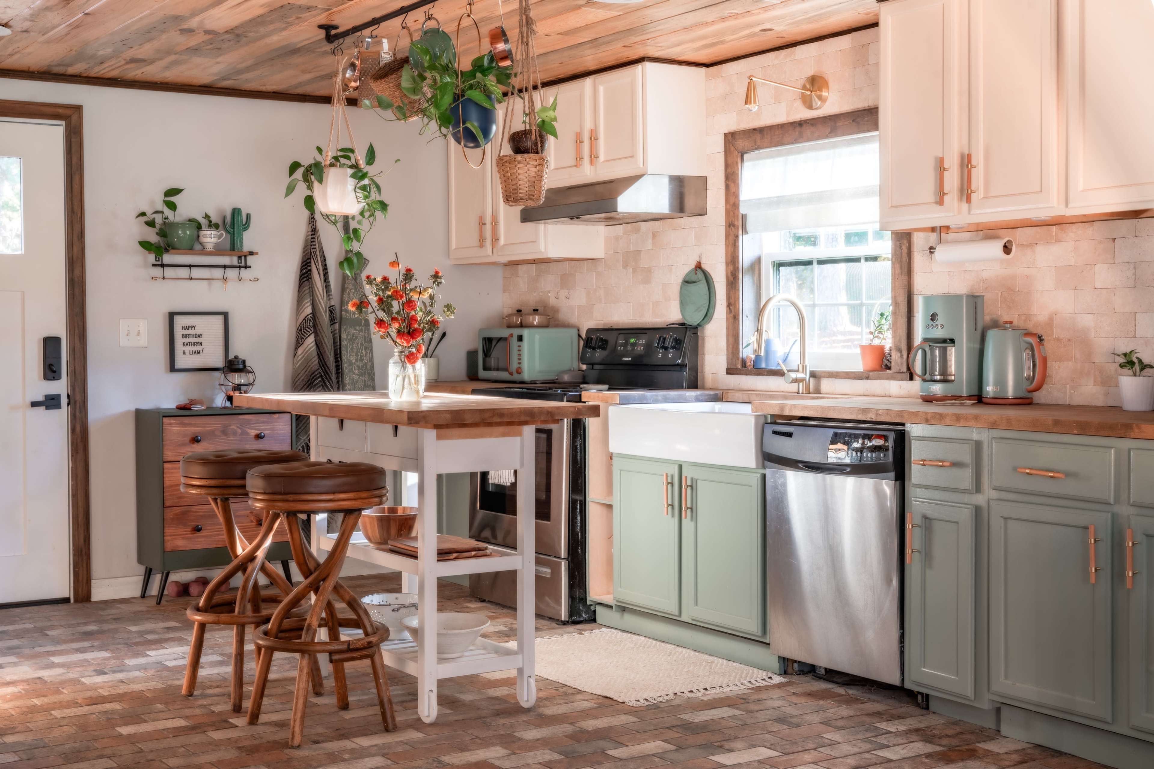 A modern kitchen features green and white cabinets, a wood countertop island with two stools, and a variety of plants hanging and displayed throughout the space.