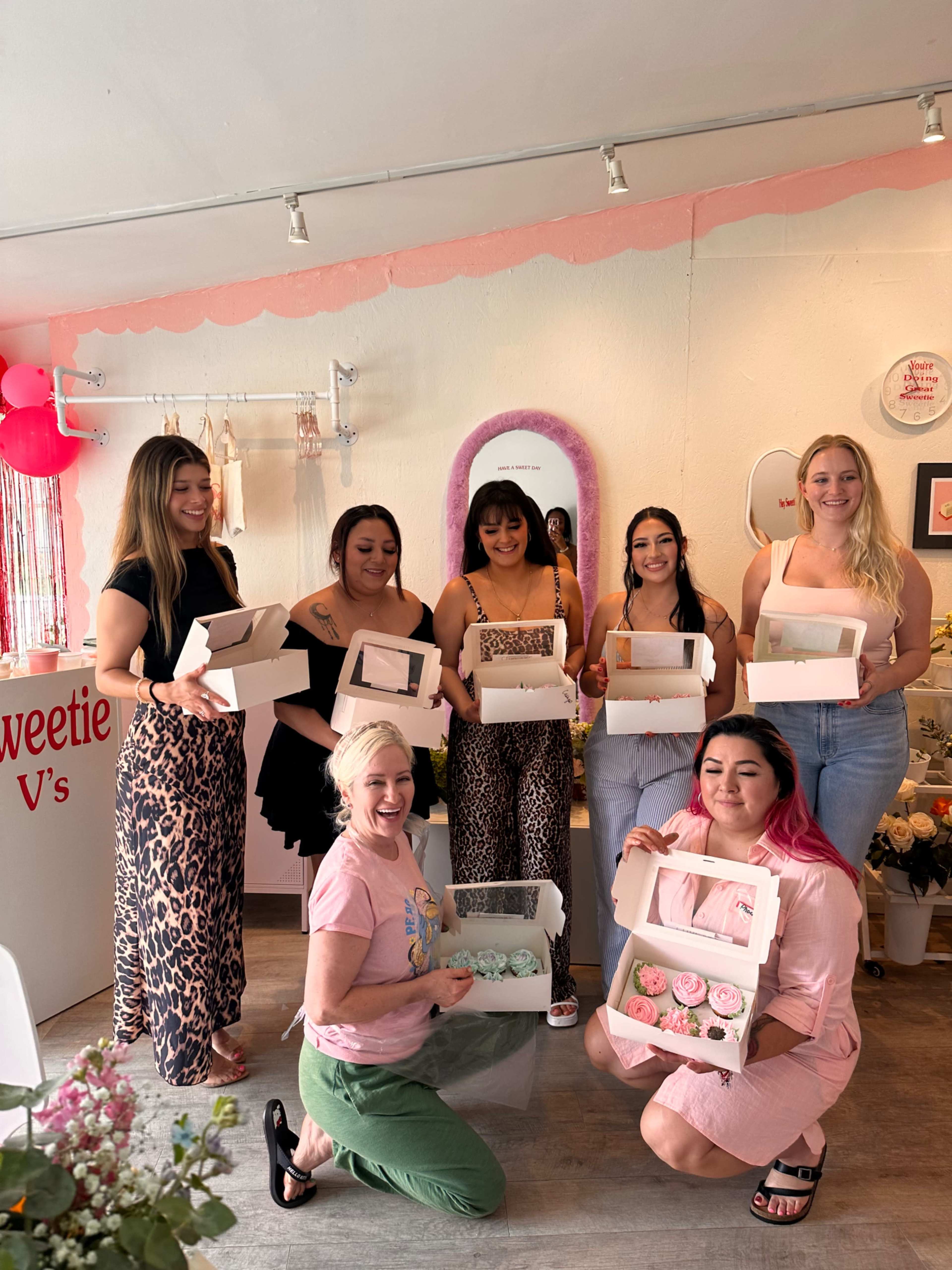 A group of seven women stands in a brightly lit shop, each holding a box of baked goods, with a display of flowers and decorations in the background.