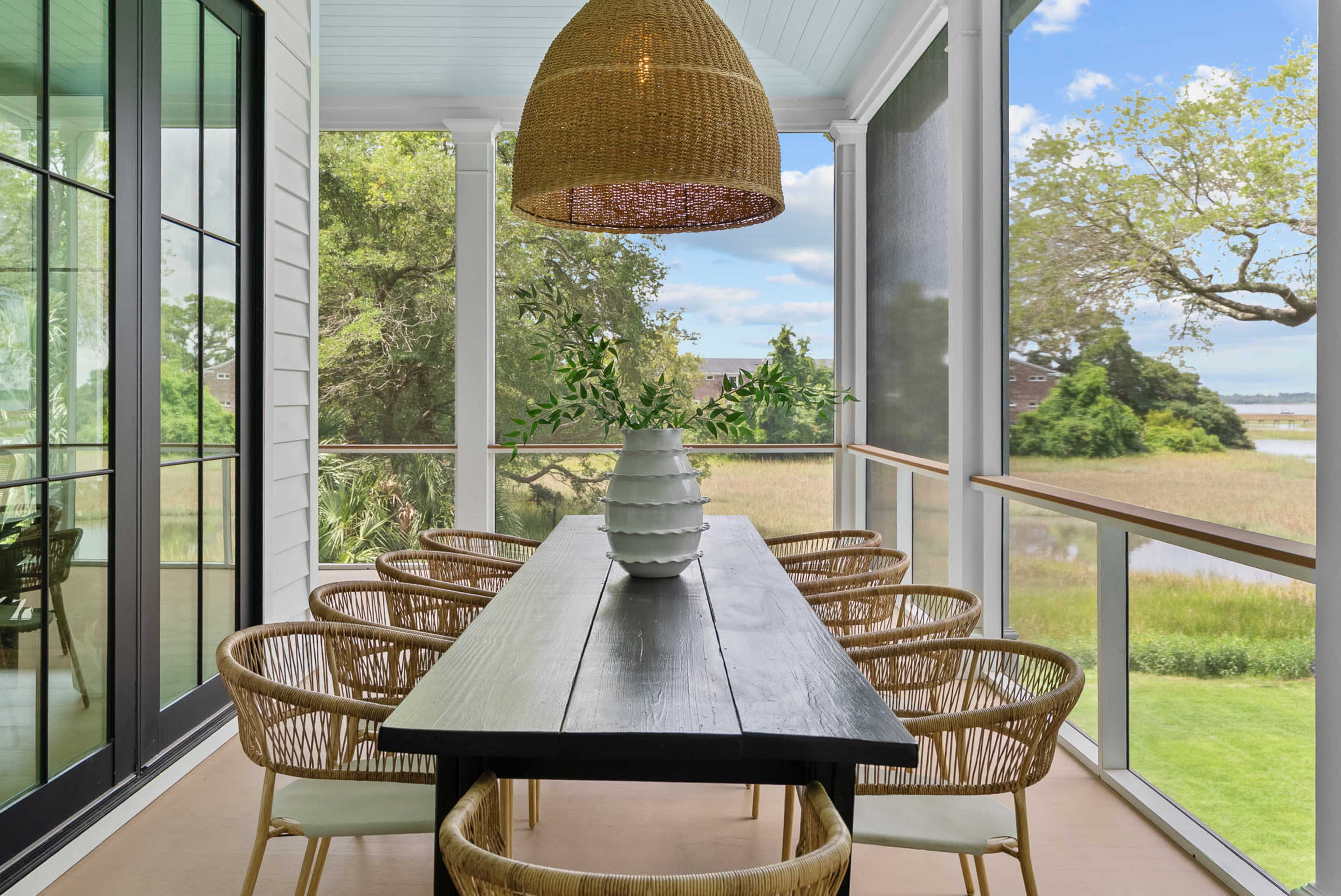 A long wooden dining table with eight rattan chairs is set on a covered porch overlooking a grassy landscape and trees.