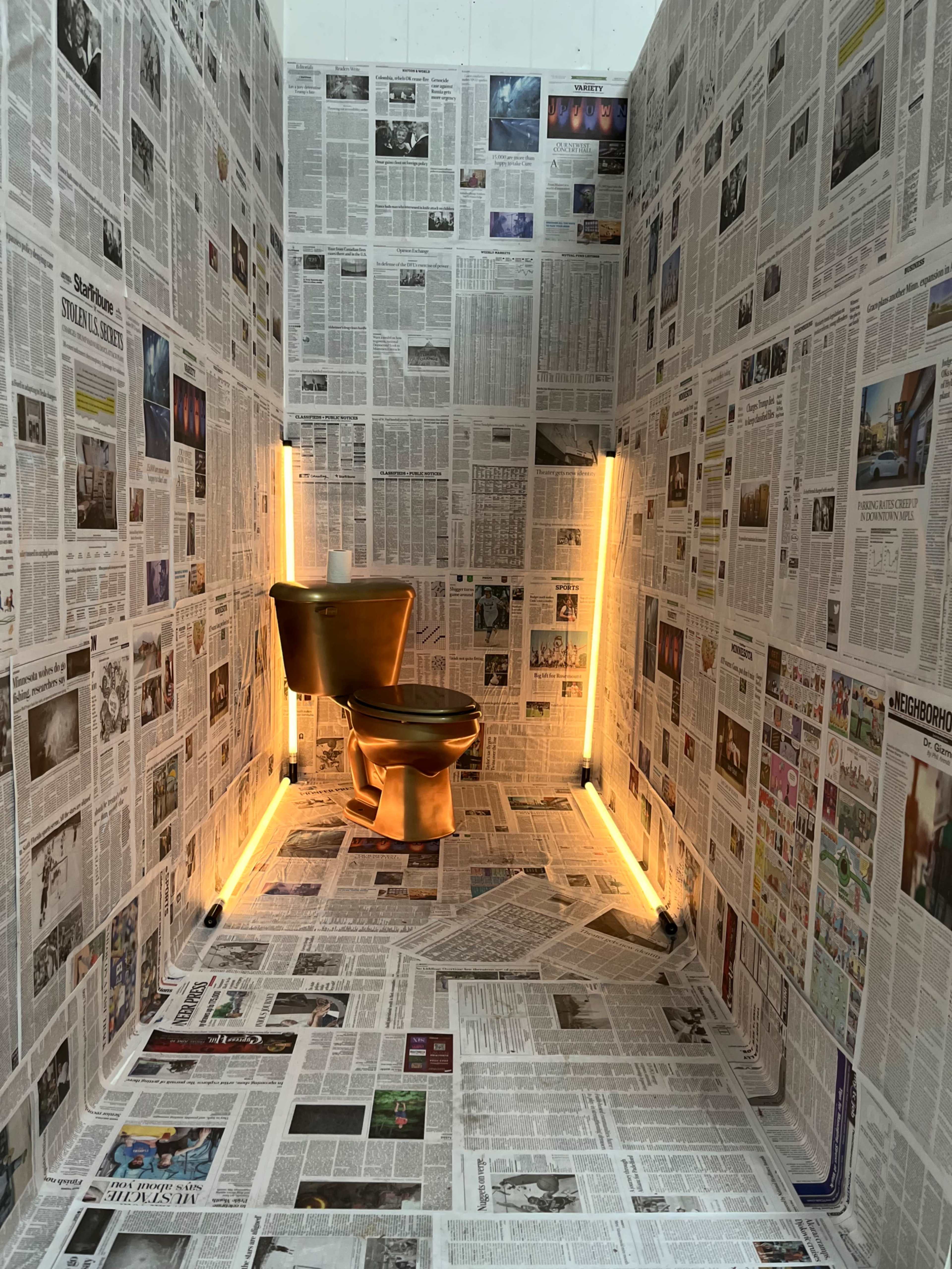 A small room entirely covered in newspapers, featuring a gold-colored toilet centered against a backdrop of newsprint with ambient lighting along the walls.