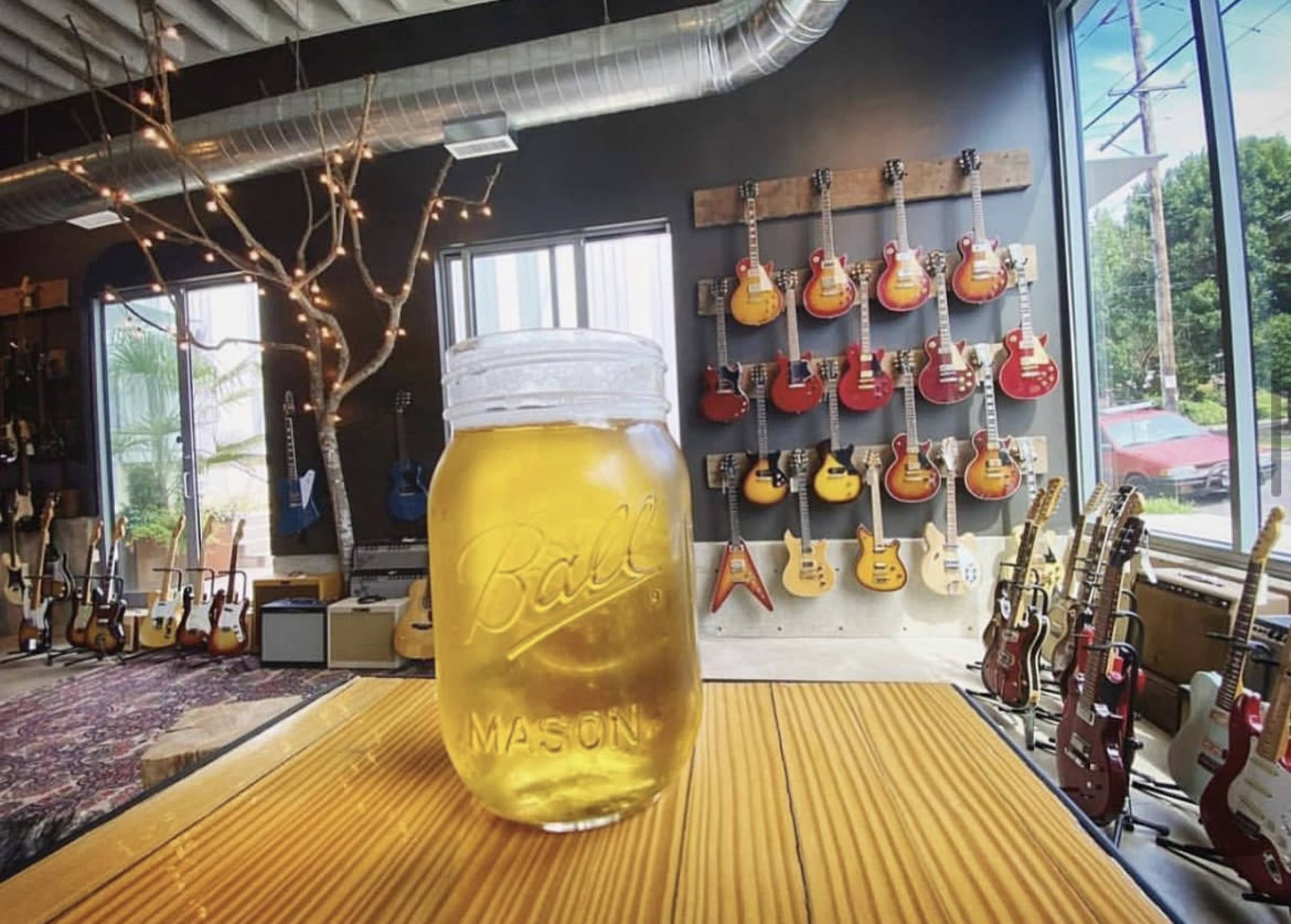 A mason jar filled with a light-colored beverage sits on a wooden table in a room with various electric guitars displayed on the walls.