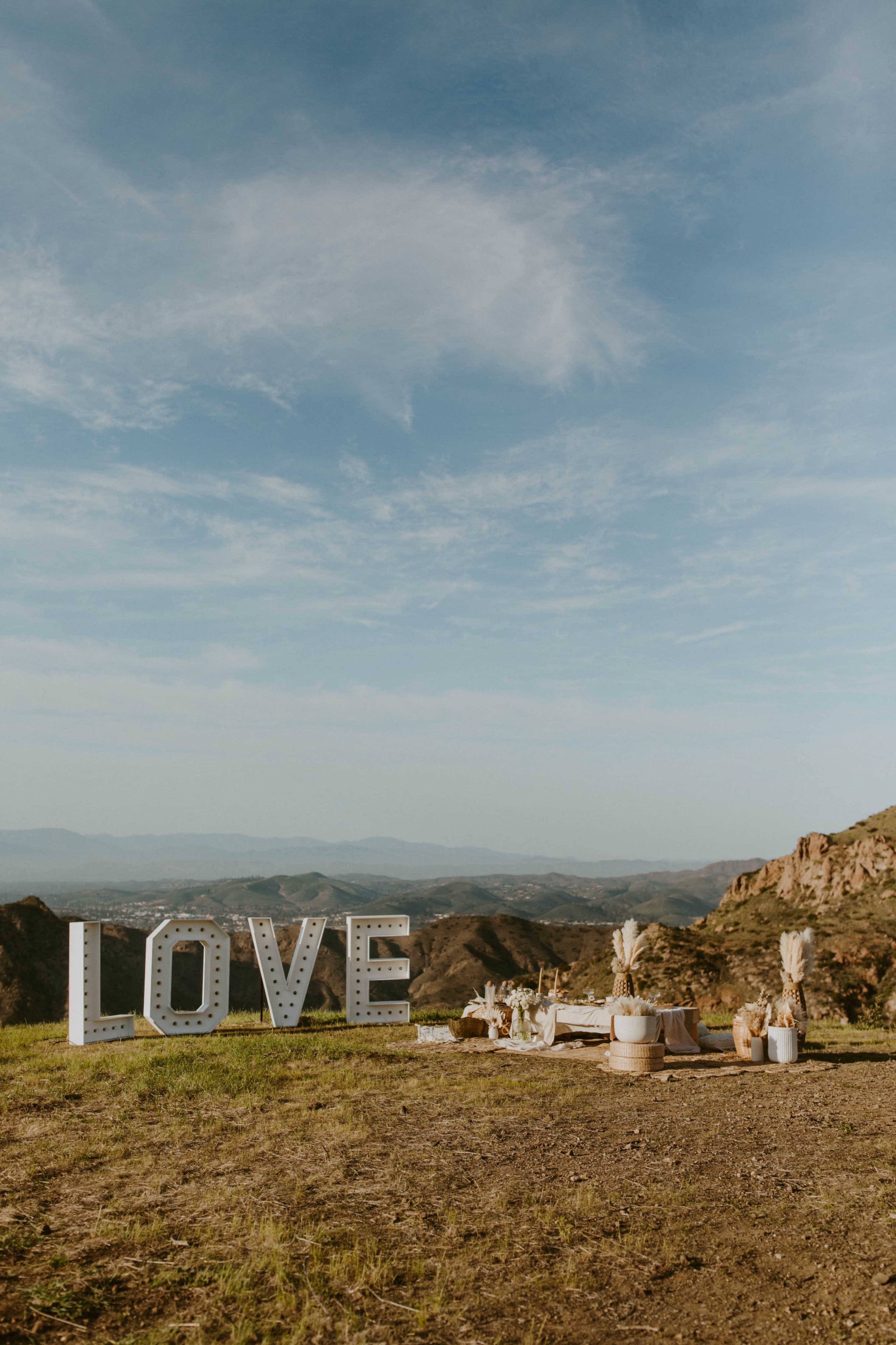 A picnic setup in a scenic outdoor location with a large illuminated "LOVE" sign and decorative elements against a backdrop of hills and a blue sky.