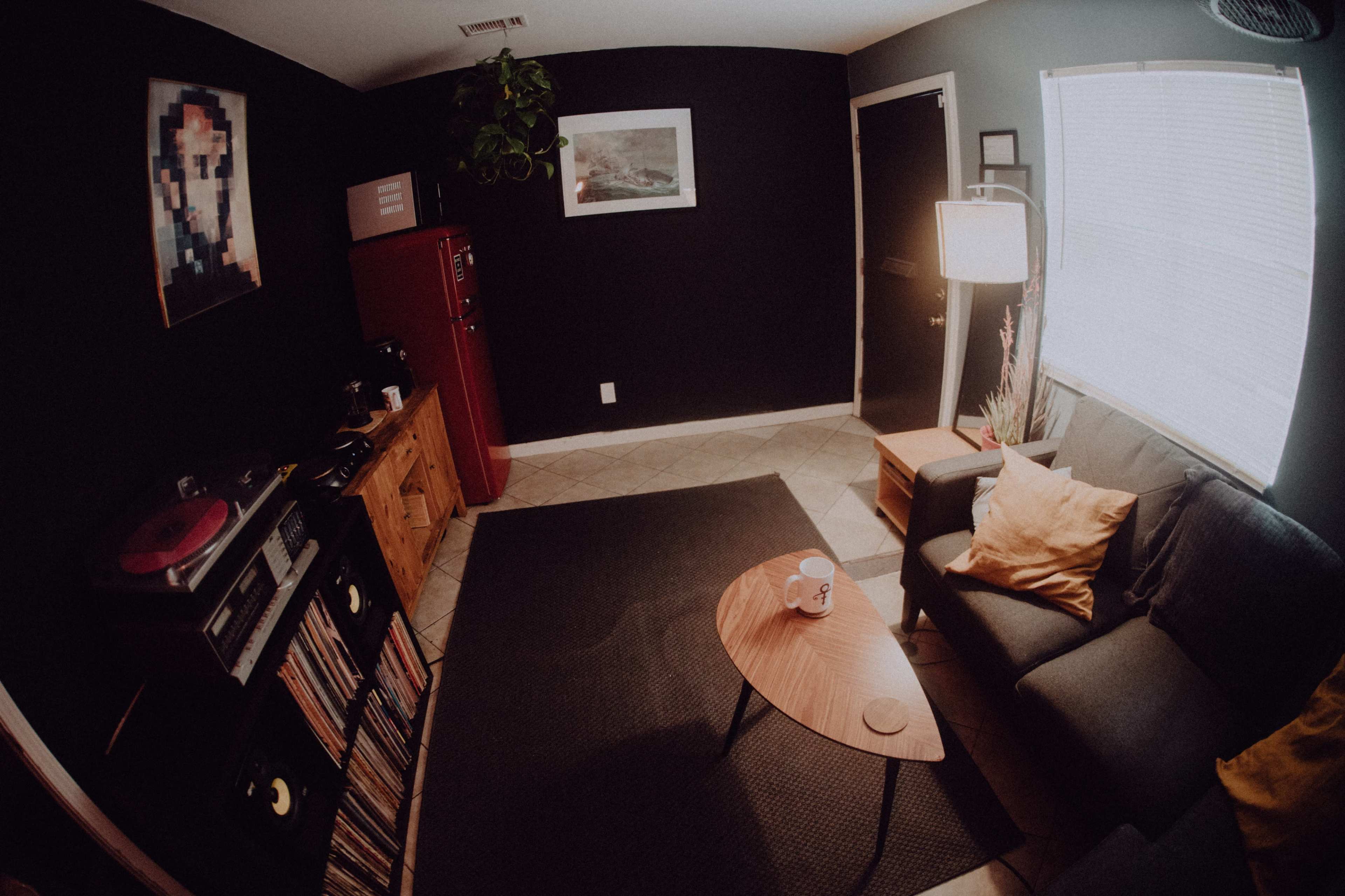 A living room features a couch, a coffee table, a vinyl record player, and a red refrigerator against dark walls.