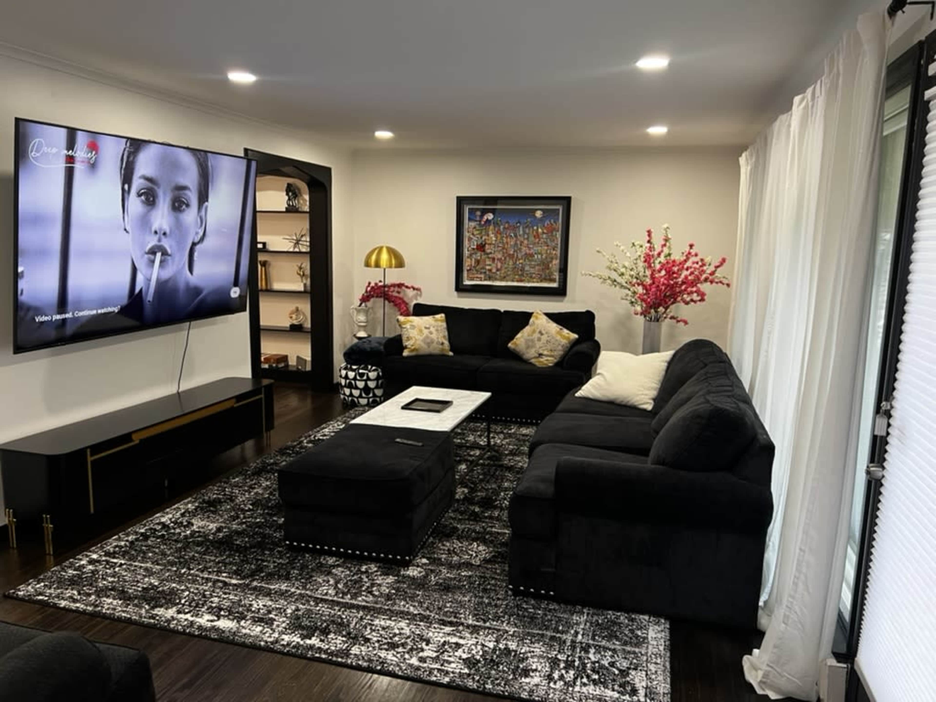 The image shows a modern living room featuring a large black sectional sofa, a coffee table, a flat-screen TV mounted on the wall, and decorative shelves with items and artwork.