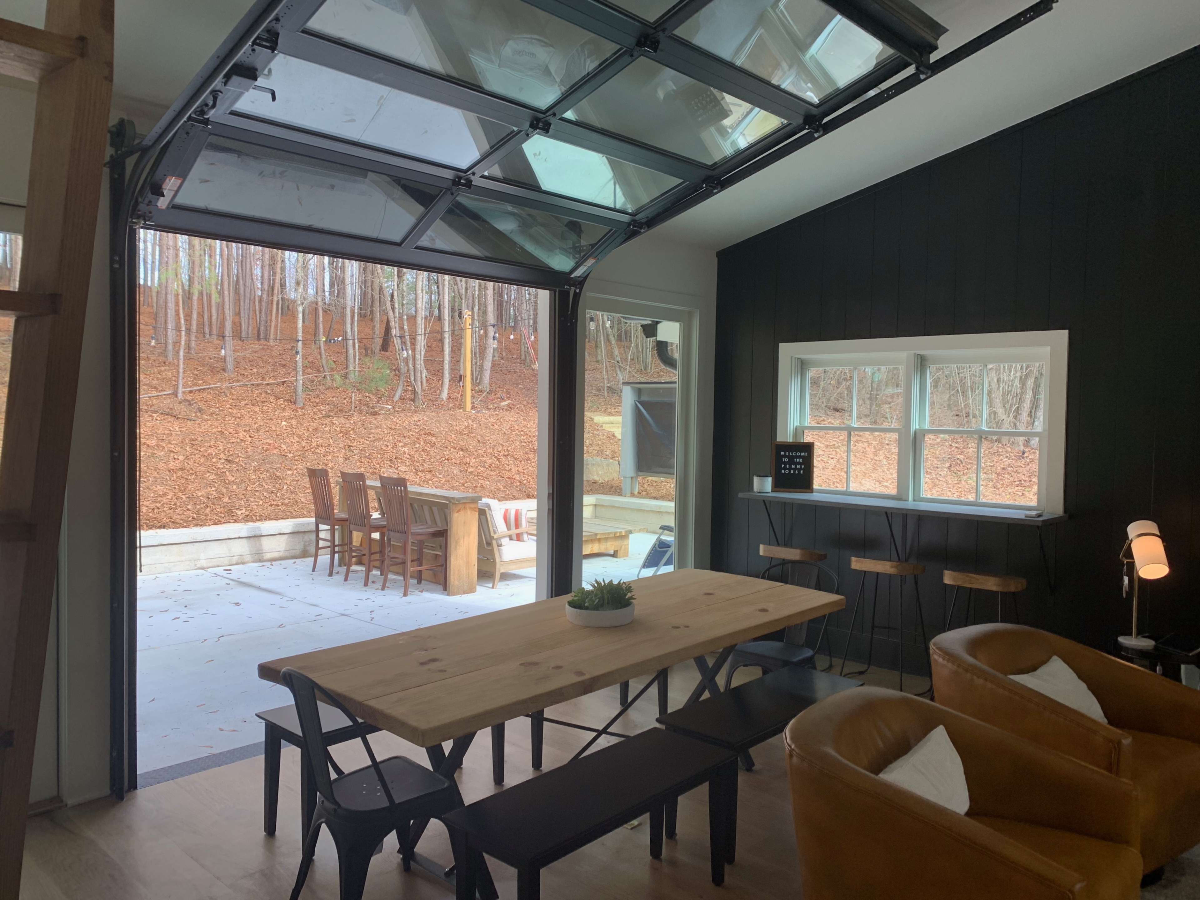A modern interior space with a wooden dining table, two leather chairs, and a garage door that opens to a patio surrounded by trees with fallen leaves.