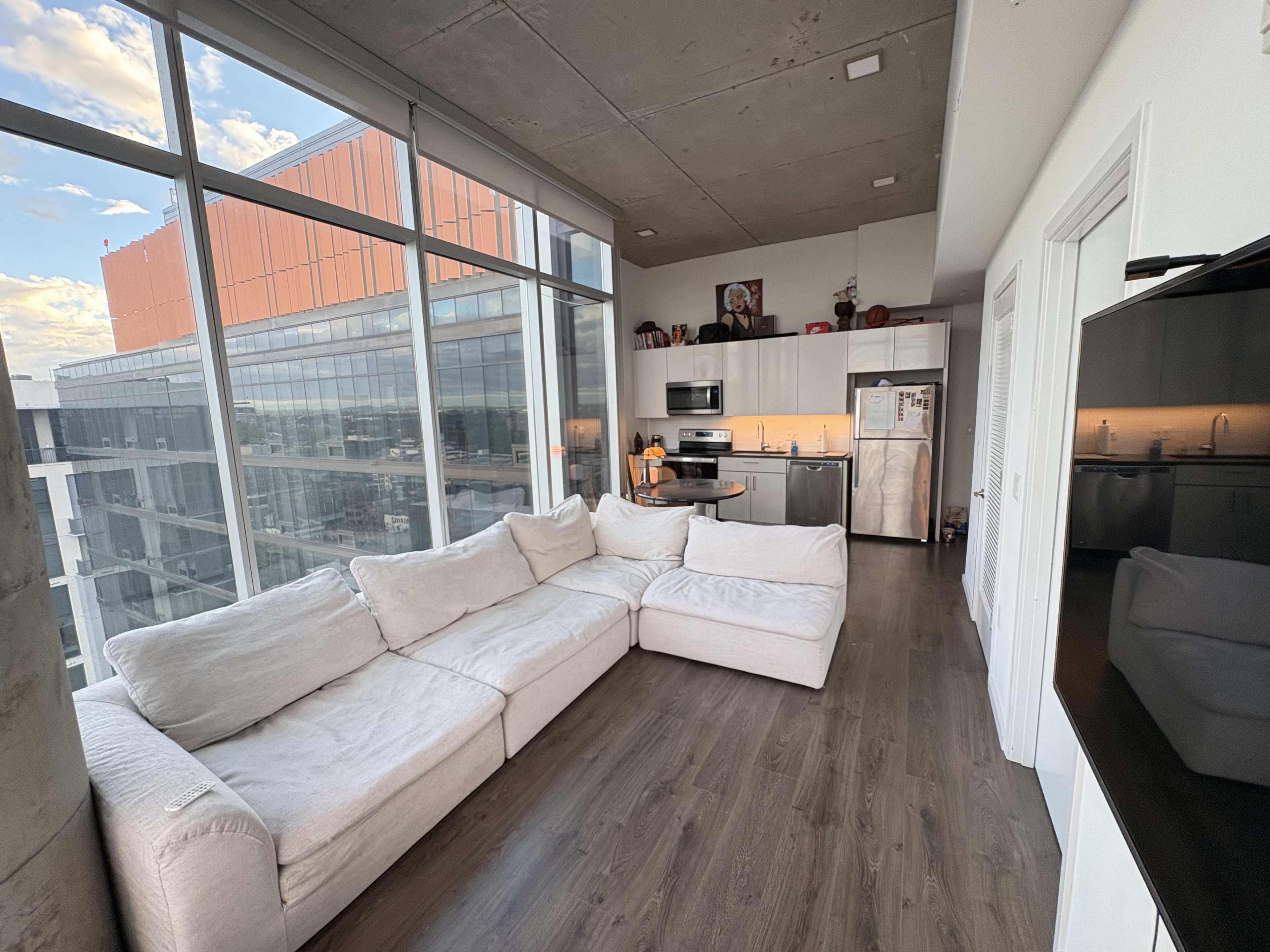 The image shows a modern, open-concept living space featuring a sectional sofa, a kitchen with stainless steel appliances, and large windows displaying an urban landscape.