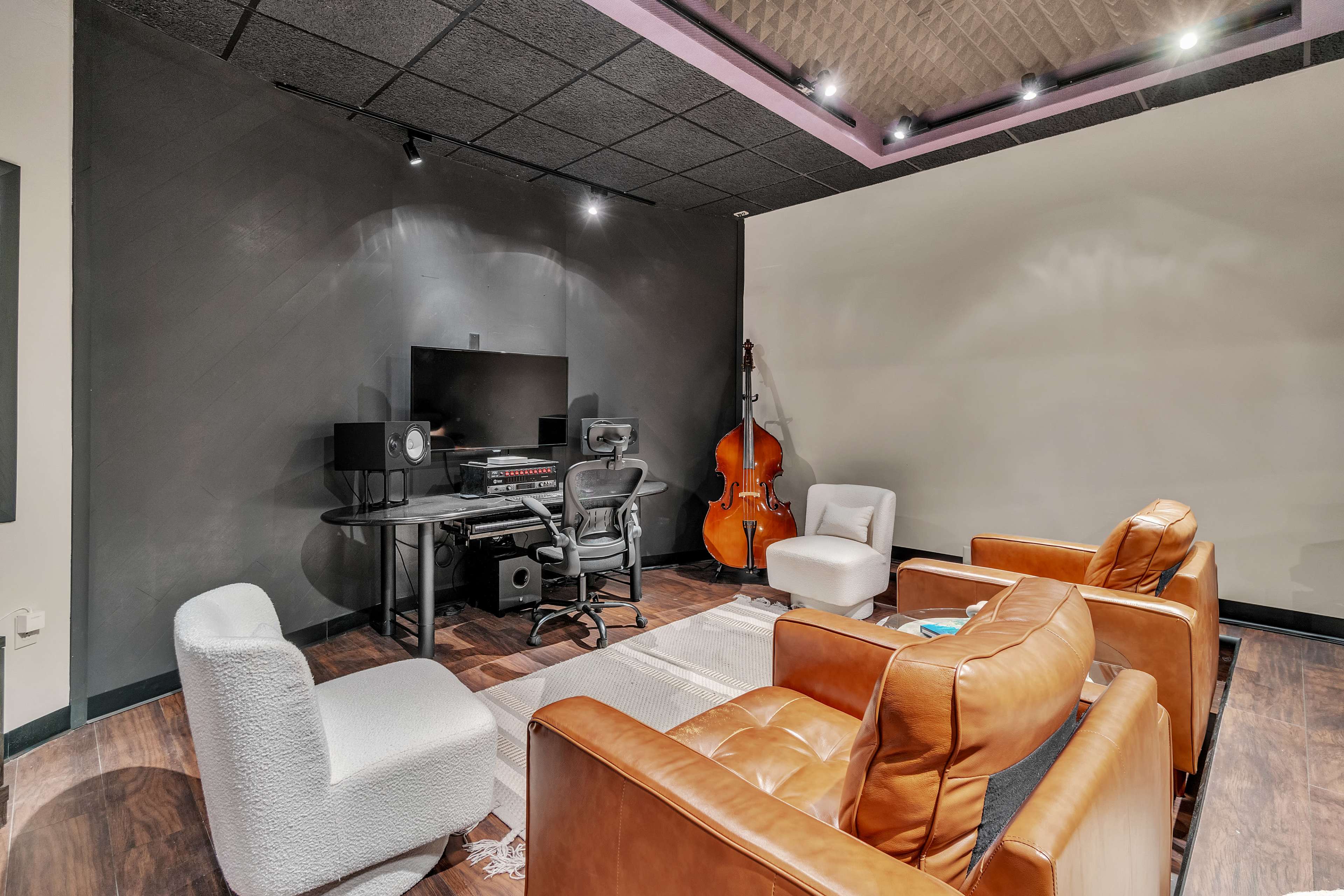 The image shows a music studio with a desk equipped for sound production, a large monitor, two chairs, a sofa, and a double bass in the corner.