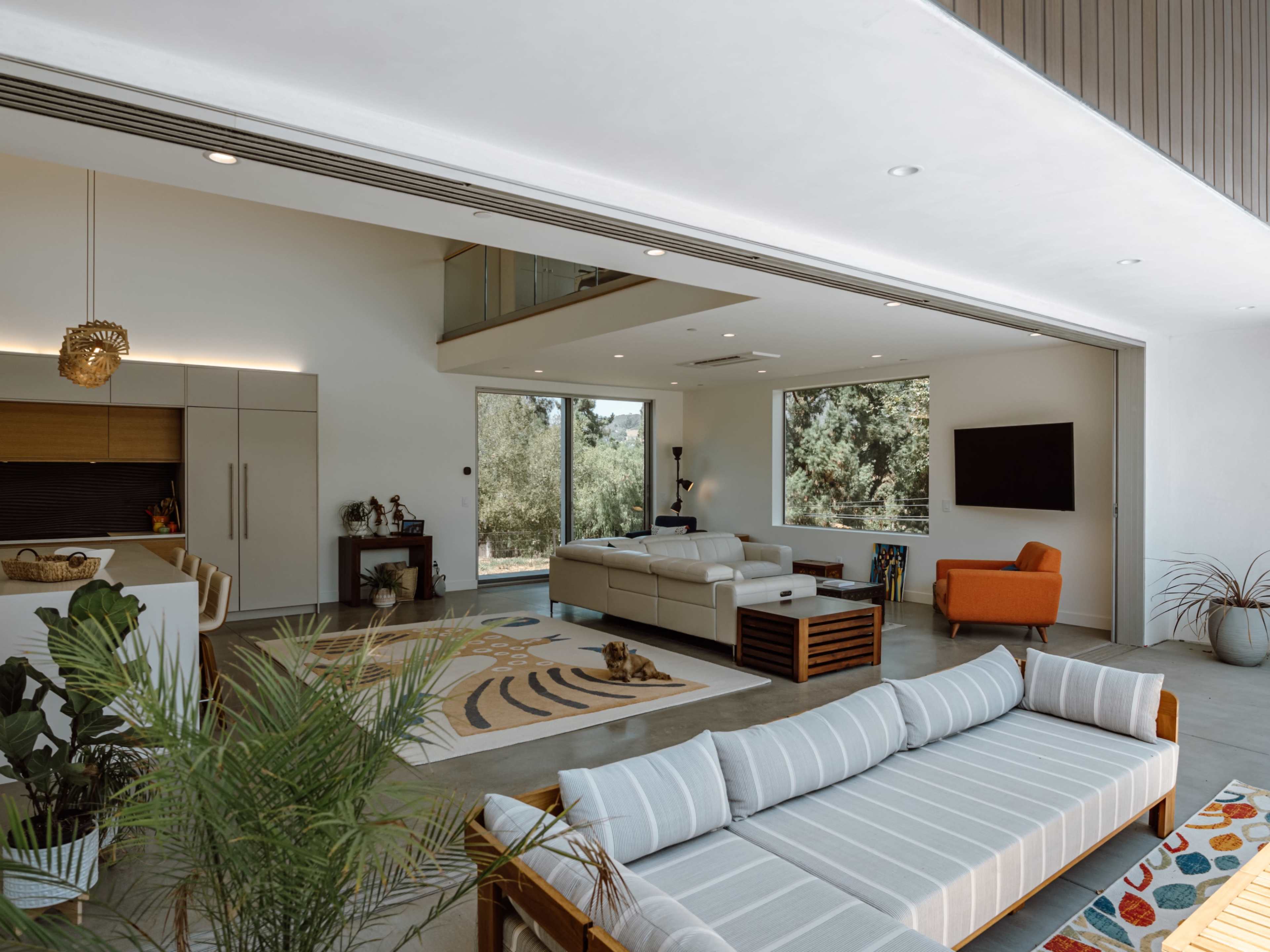 The image shows a modern living room with a spacious layout, featuring a sectional sofa, an armchair, a coffee table, and large windows that provide natural light.