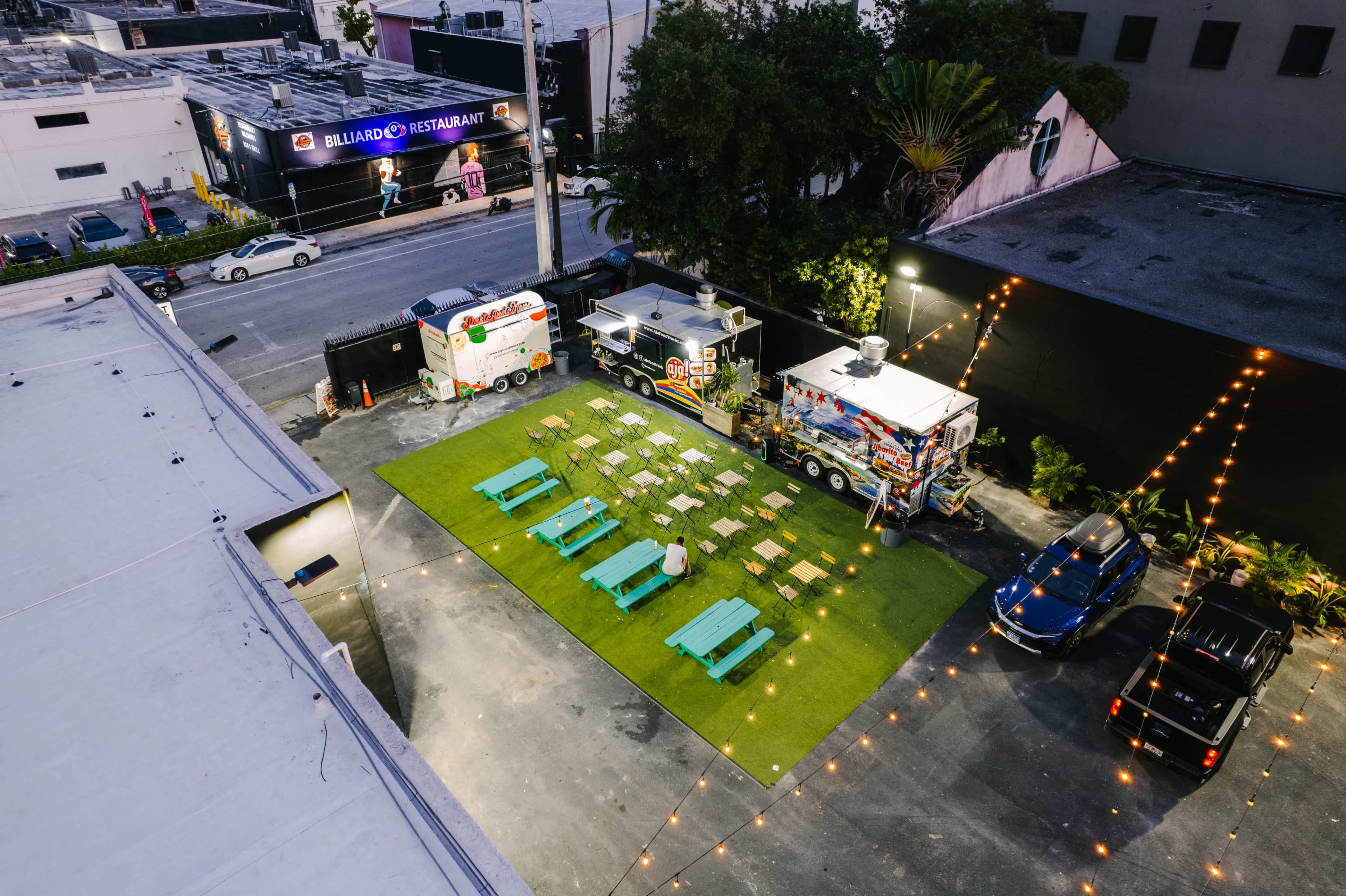 Aerial view of a food truck area featuring green seating on turf, surrounded by string lights and parked cars.