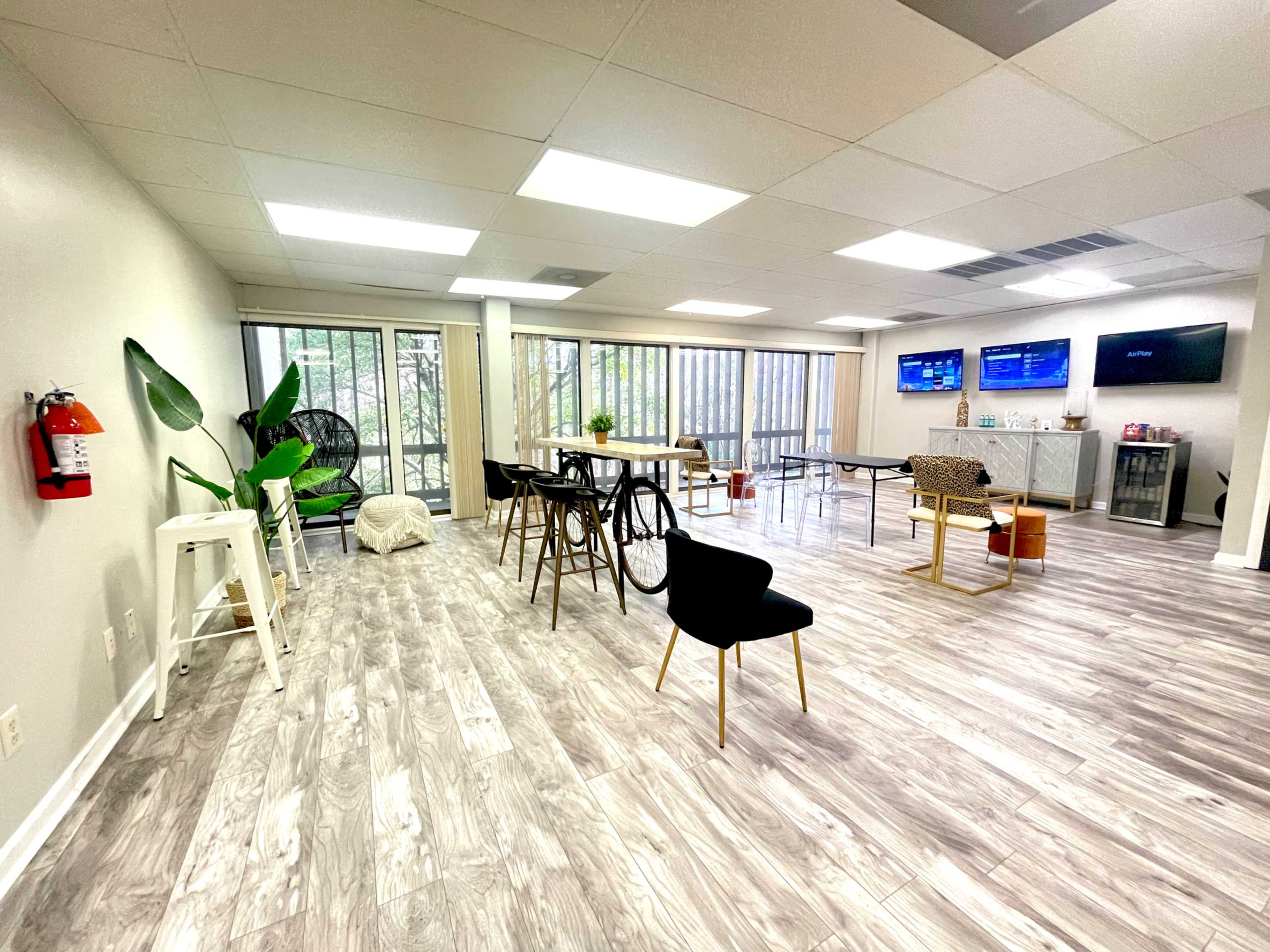 A spacious, well-lit room with multiple tables, chairs, a large window with greenery outside, and several wall-mounted displays.