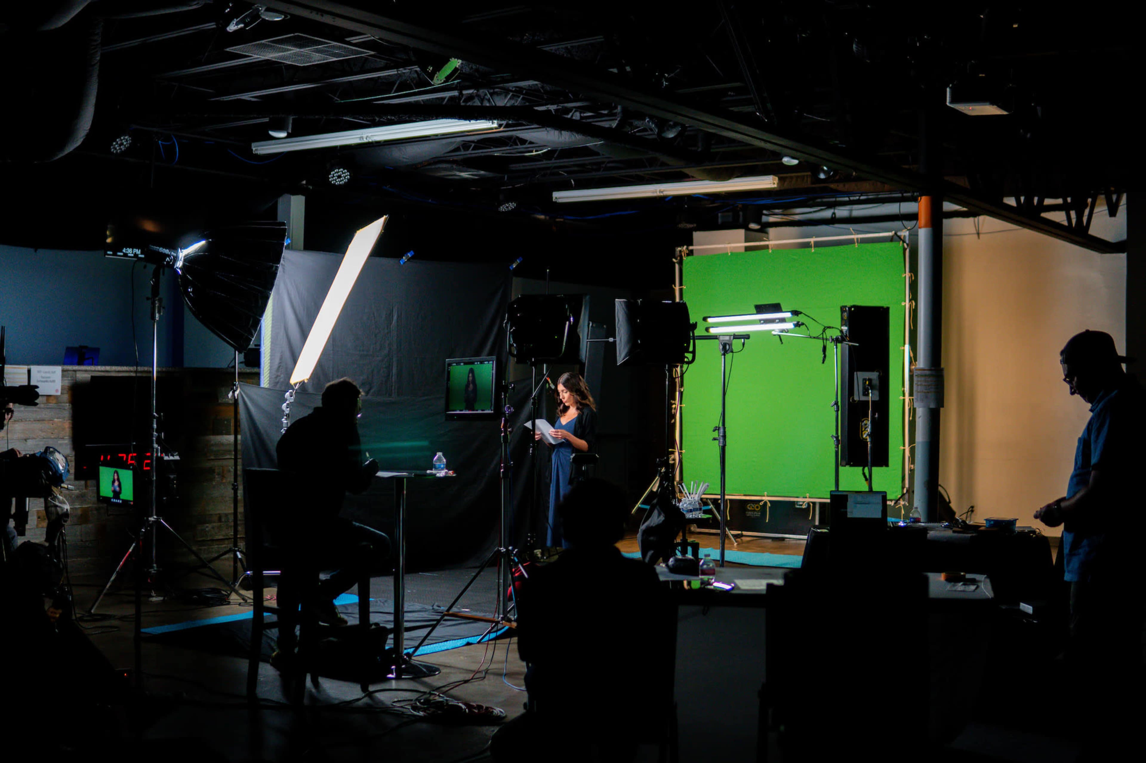 A woman stands in front of a green screen while a camera crew and equipment are set up in a studio environment.