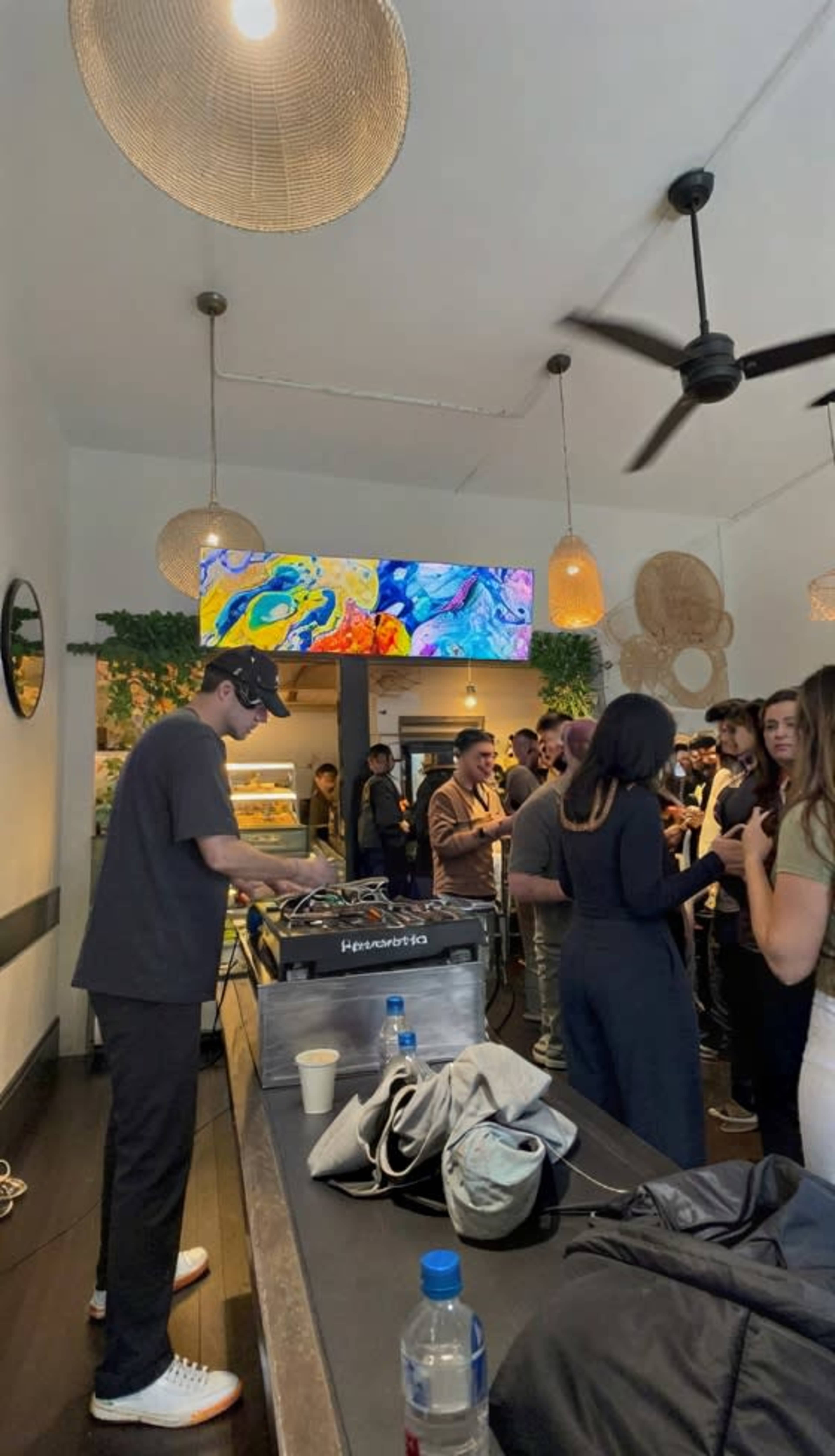 A DJ stands behind a turntable while a crowd socializes in a stylish, modern indoor space with decorative lighting and greenery.