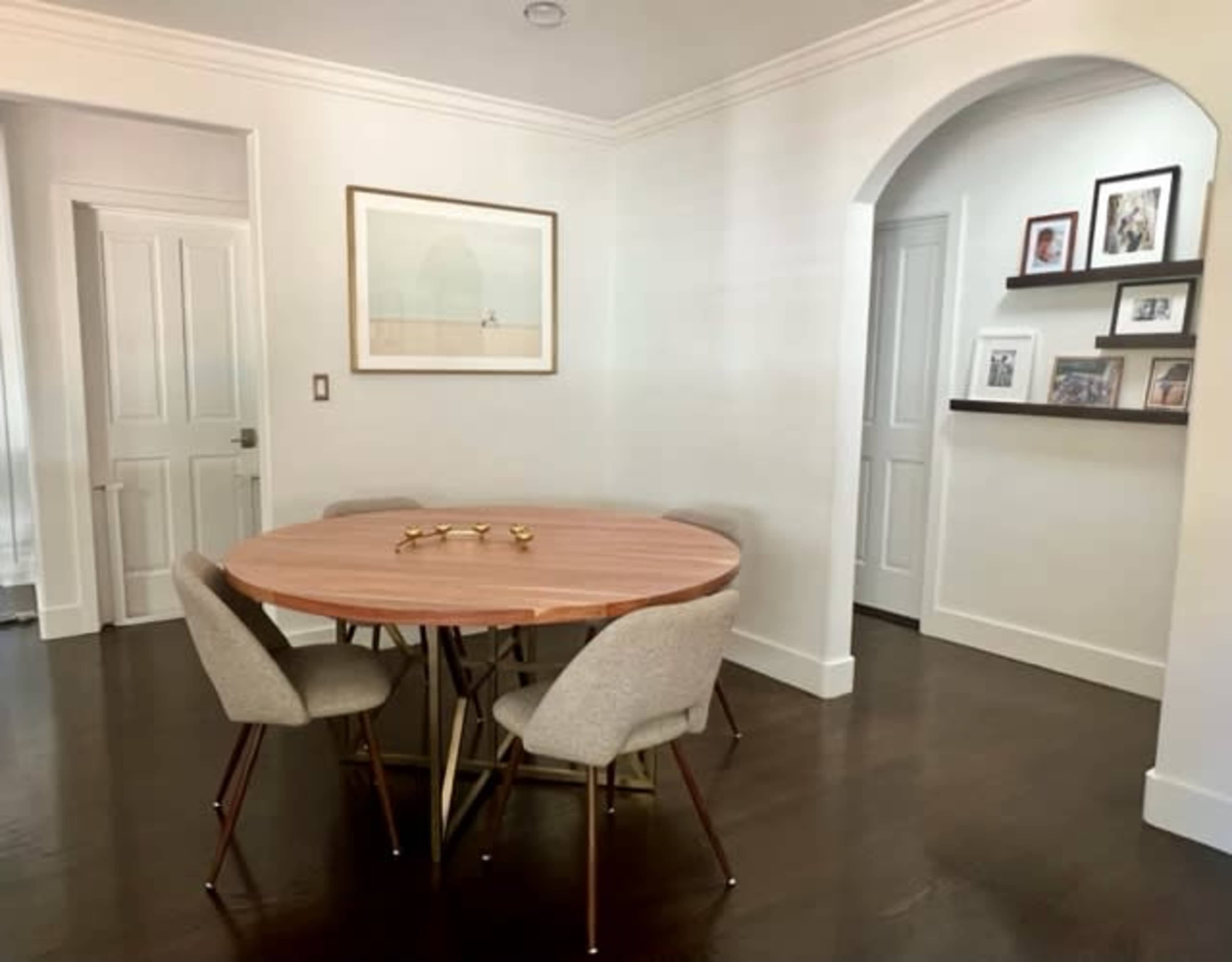 A round wooden dining table with four upholstered chairs is set in a brightly lit room with light-colored walls and a doorway leading to another area.