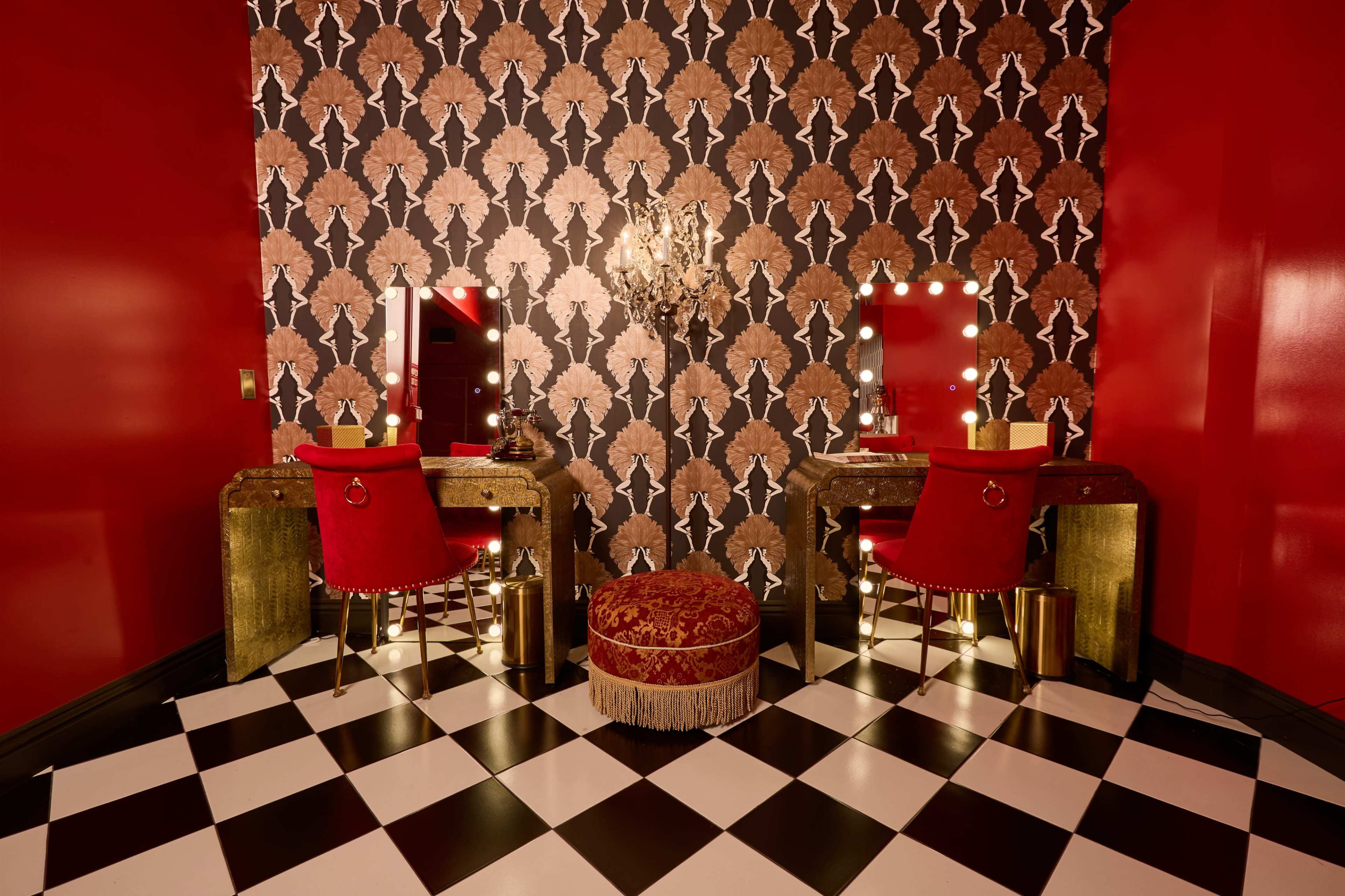 The image features a room with a bold red and black patterned wallpaper, two mirrored vanities with red chairs, a chandelier, and a checkered floor.