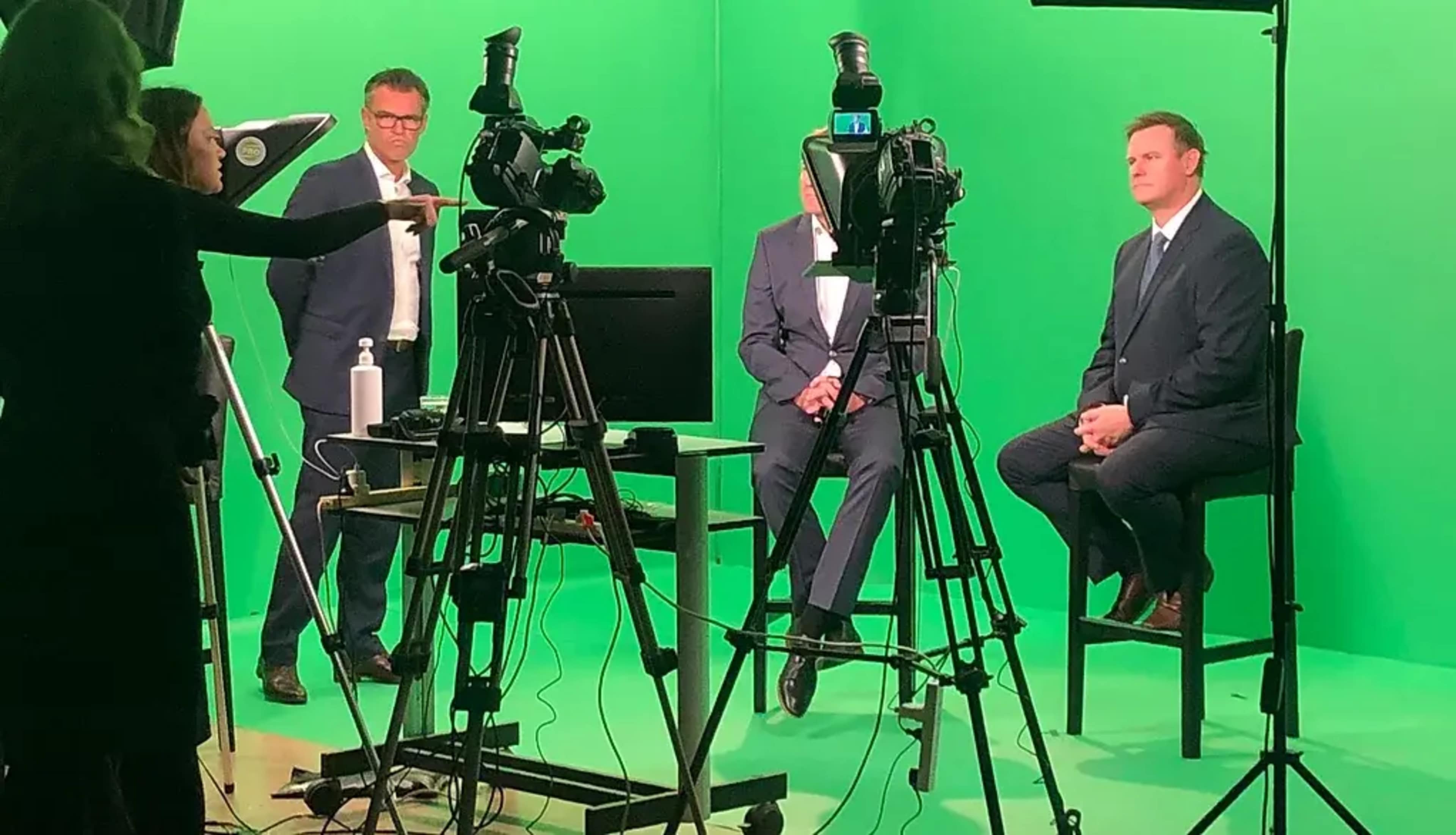 Two men in suits sit on stools in front of a green screen while a person gestures towards a camera setup.