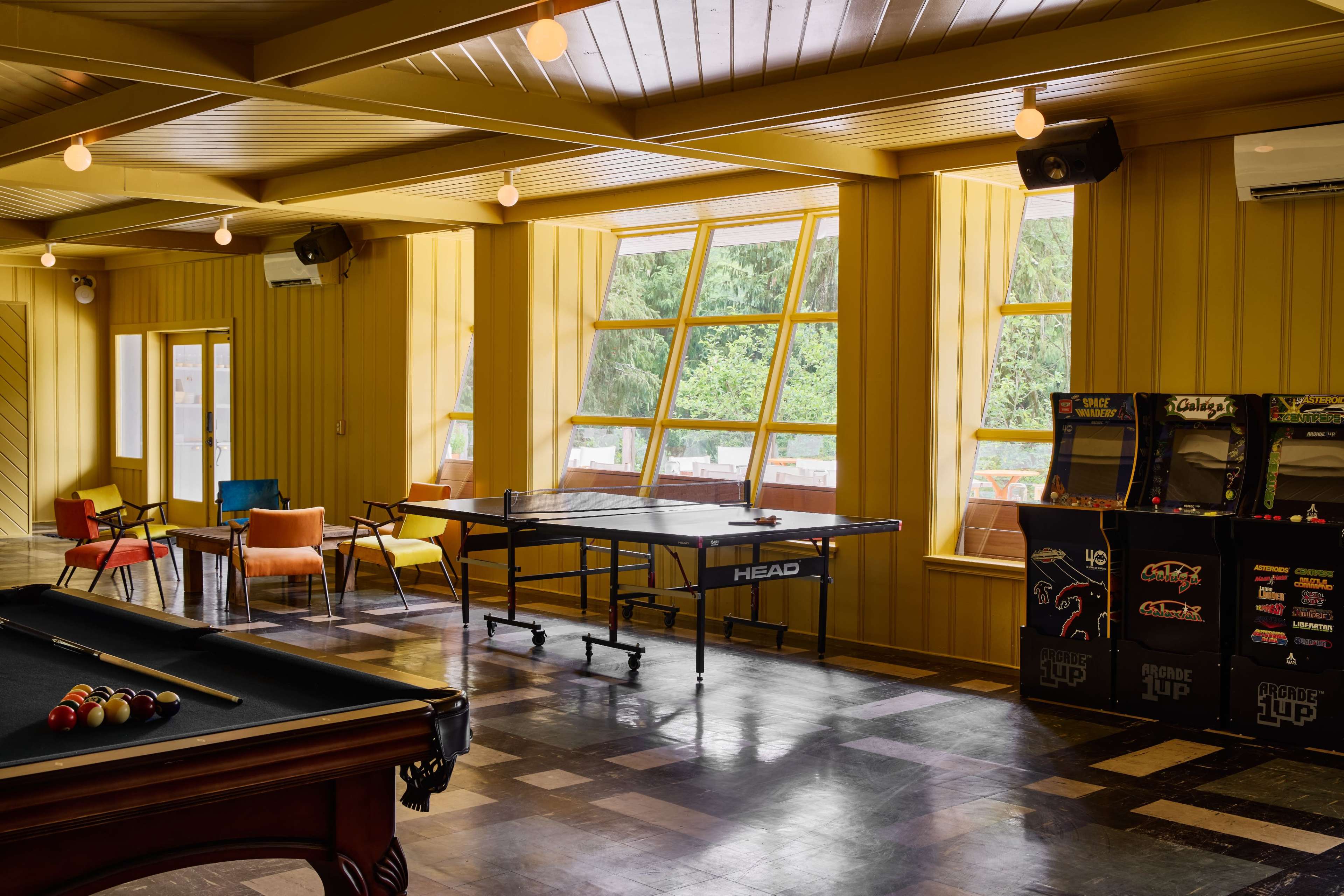 A recreational room features a pool table, a ping pong table by large windows, and arcade machines against a yellow-paneled wall.