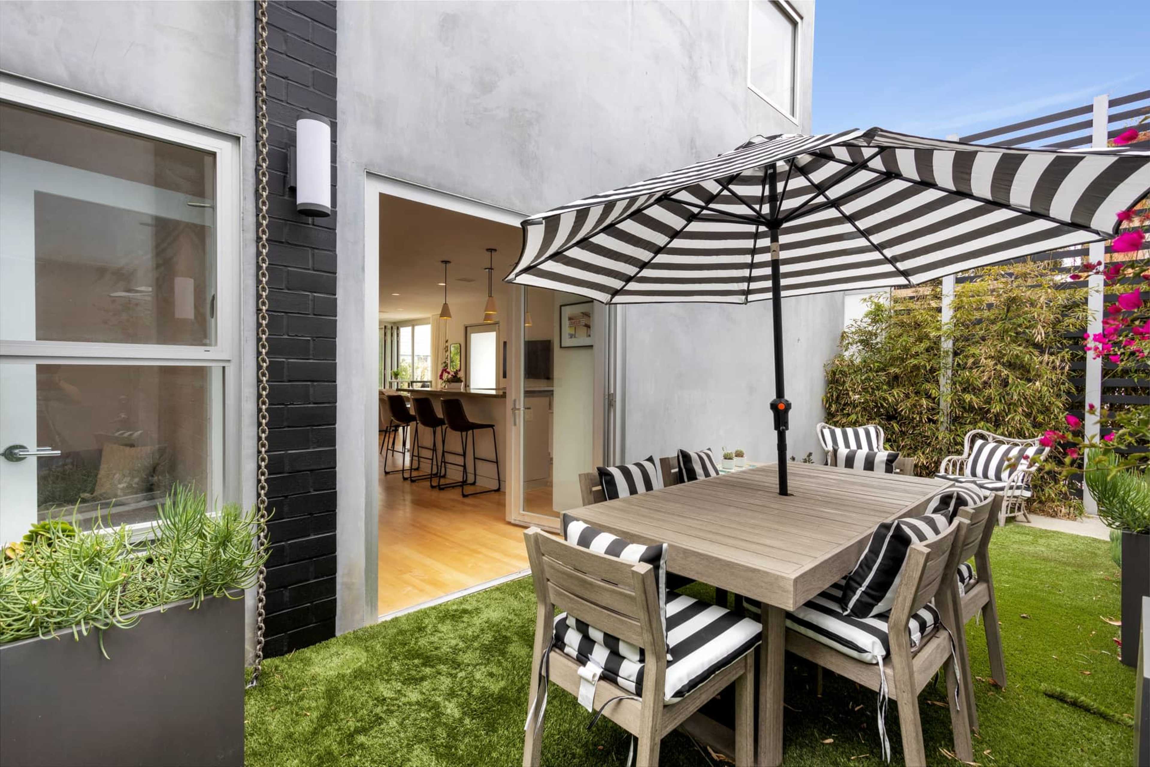 A patio area features a striped umbrella over a wooden dining table surrounded by chairs, with an entrance leading to a modern kitchen in the background.