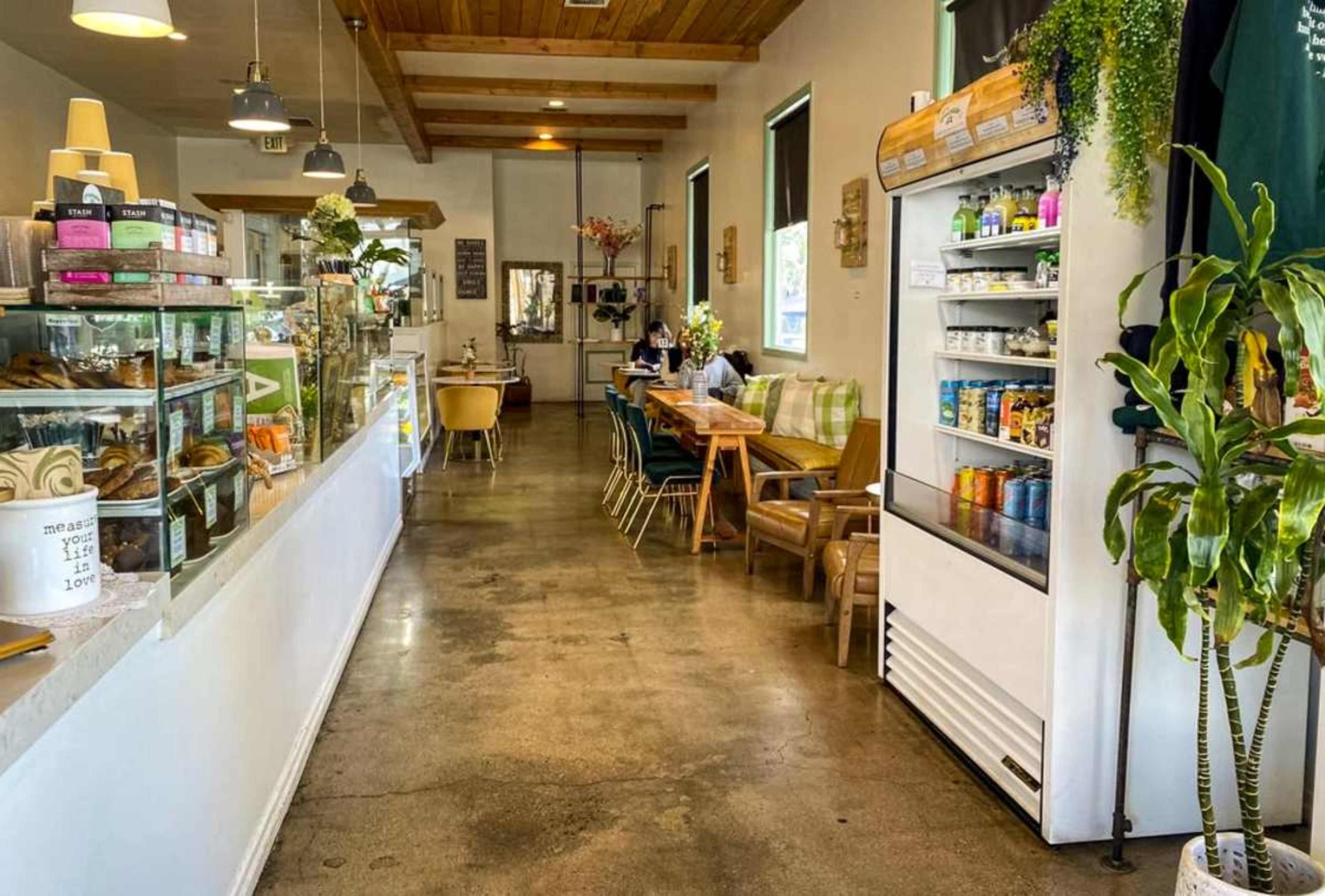 The image shows an interior of a cafe with a counter displaying pastries and a seating area featuring tables and chairs along a long corridor.