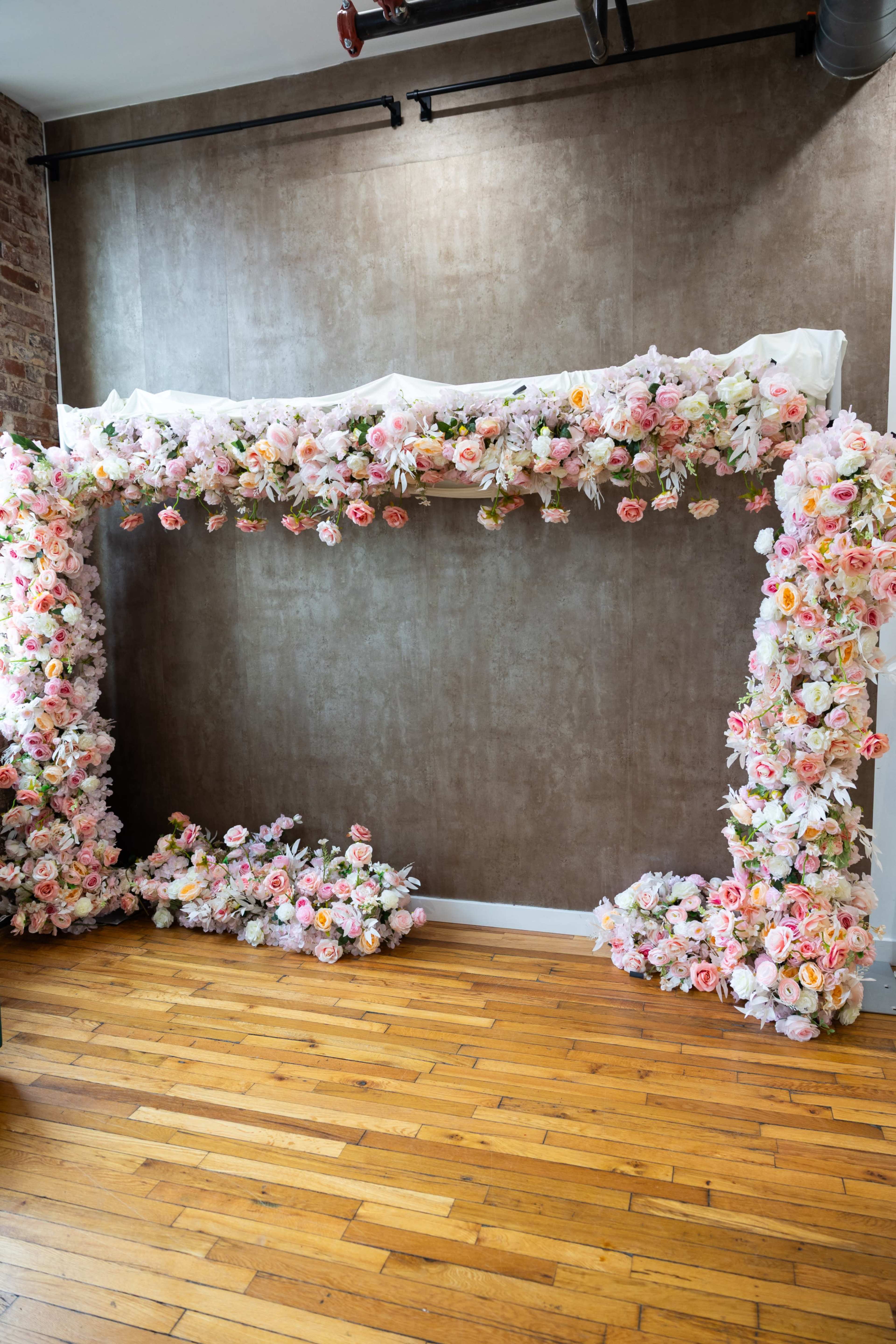 A large floral arch made of pink and white flowers is set against a textured brown wall, with wooden flooring visible beneath it.
