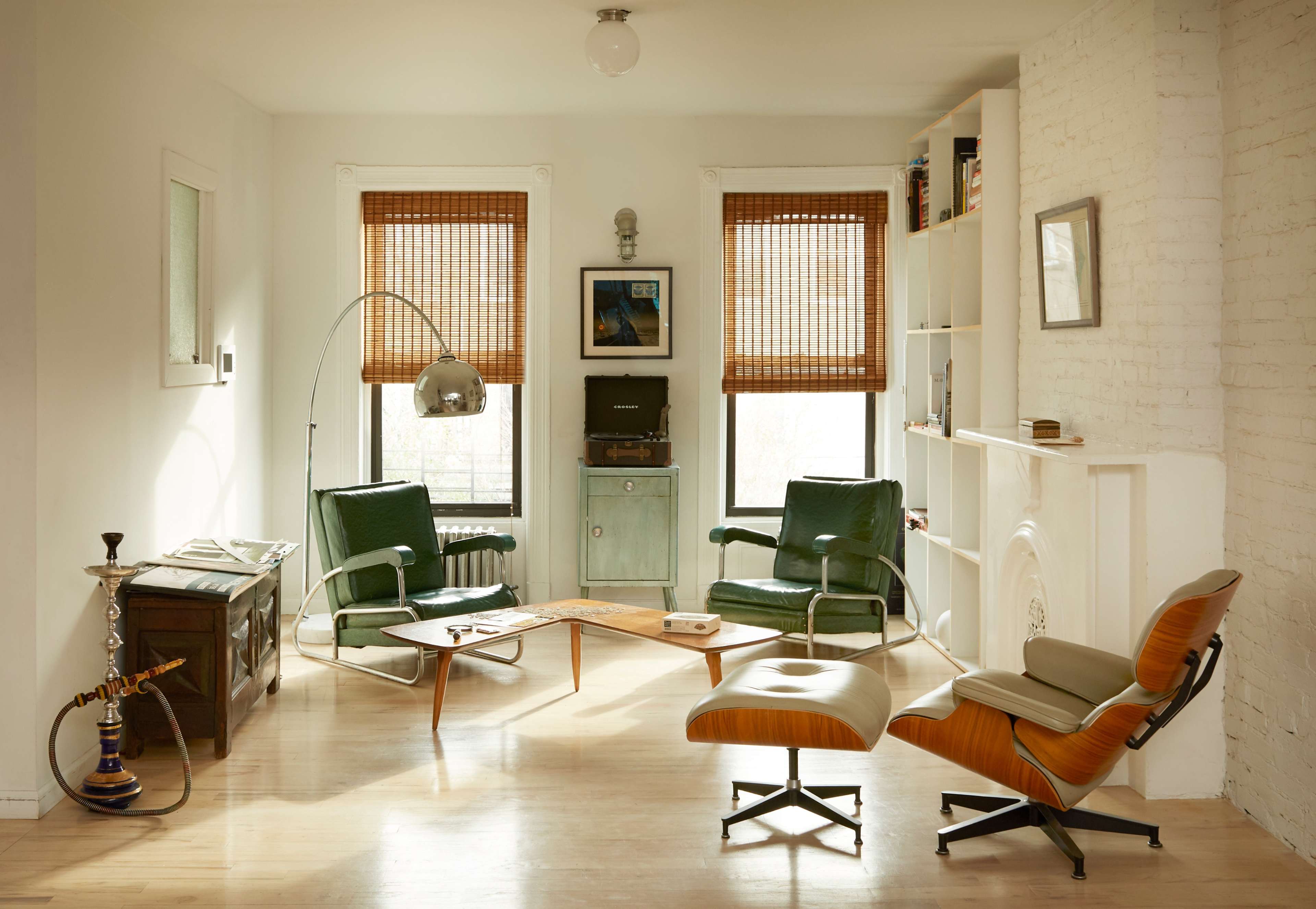 A well-lit living room features two green chairs, a wooden coffee table, and a vintage record player against a backdrop of white walls and natural light from window shades.
