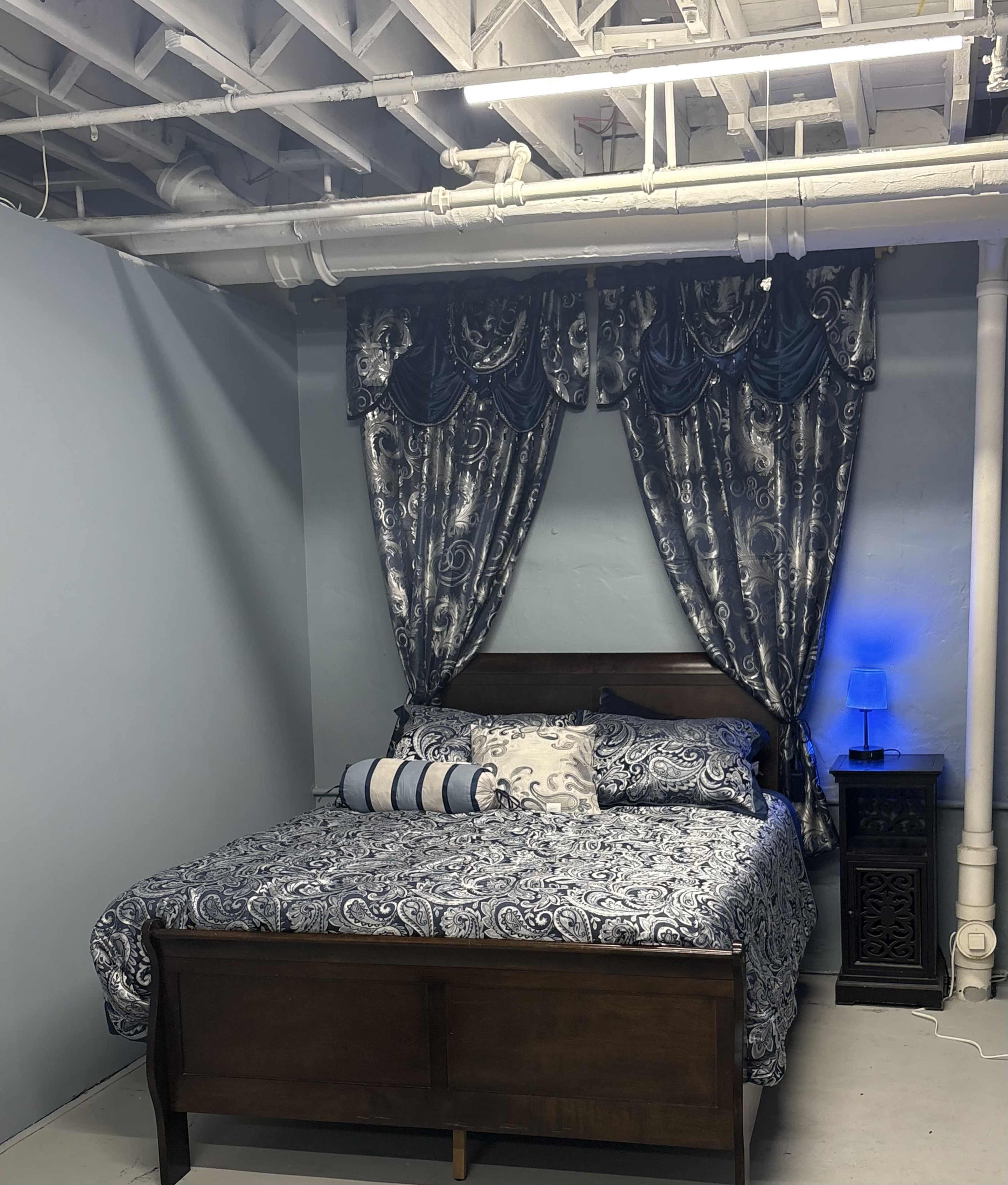 The image shows a bedroom featuring a wooden bed with a blue and white patterned duvet and matching curtains, accompanied by a nightstand and a blue lamp.