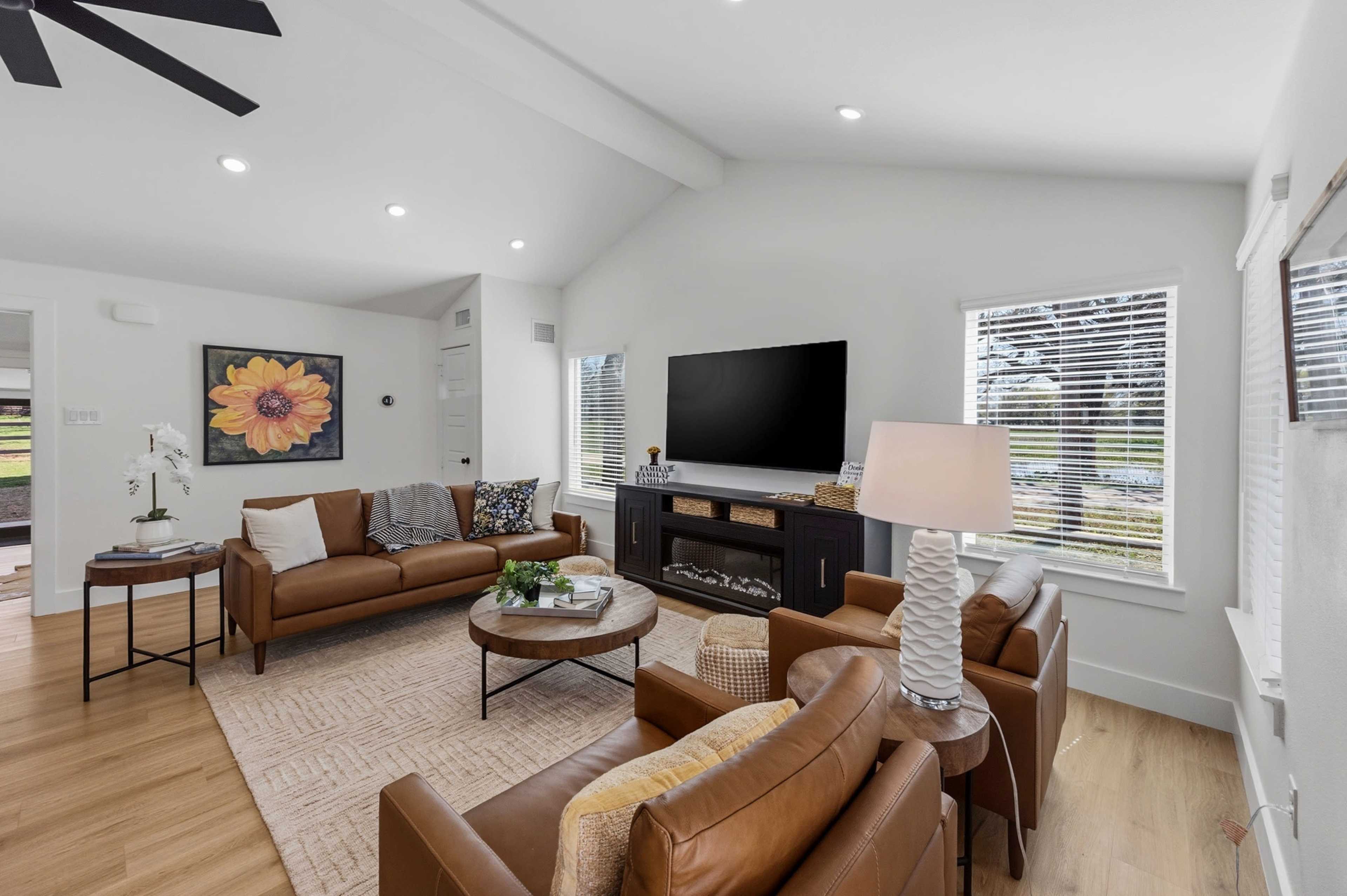 The living room features two brown leather sofas, a round coffee table, and a large television mounted on a white wall, complemented by natural light from the windows.