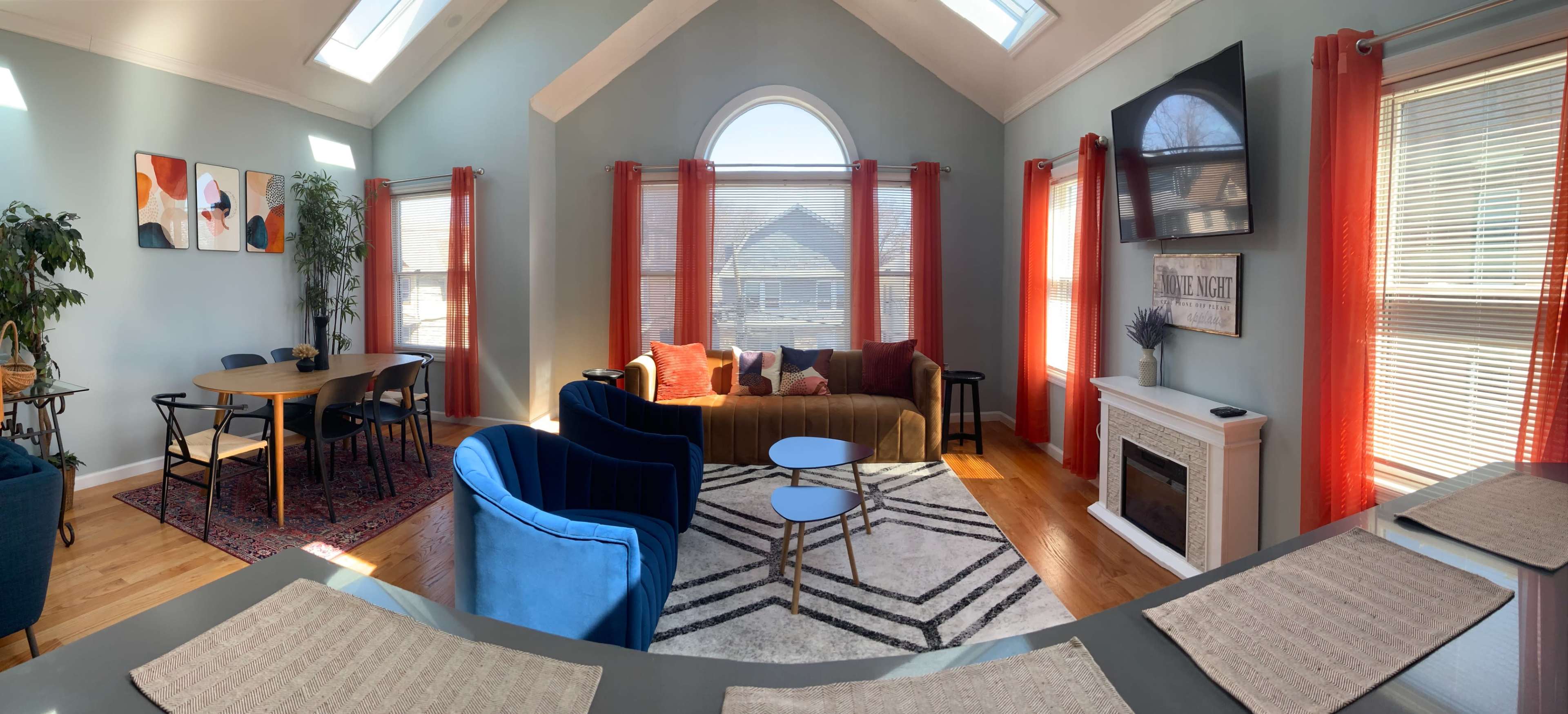 A bright living room with large windows, a brown sofa, a blue armchair, and a dining table near a fireplace.