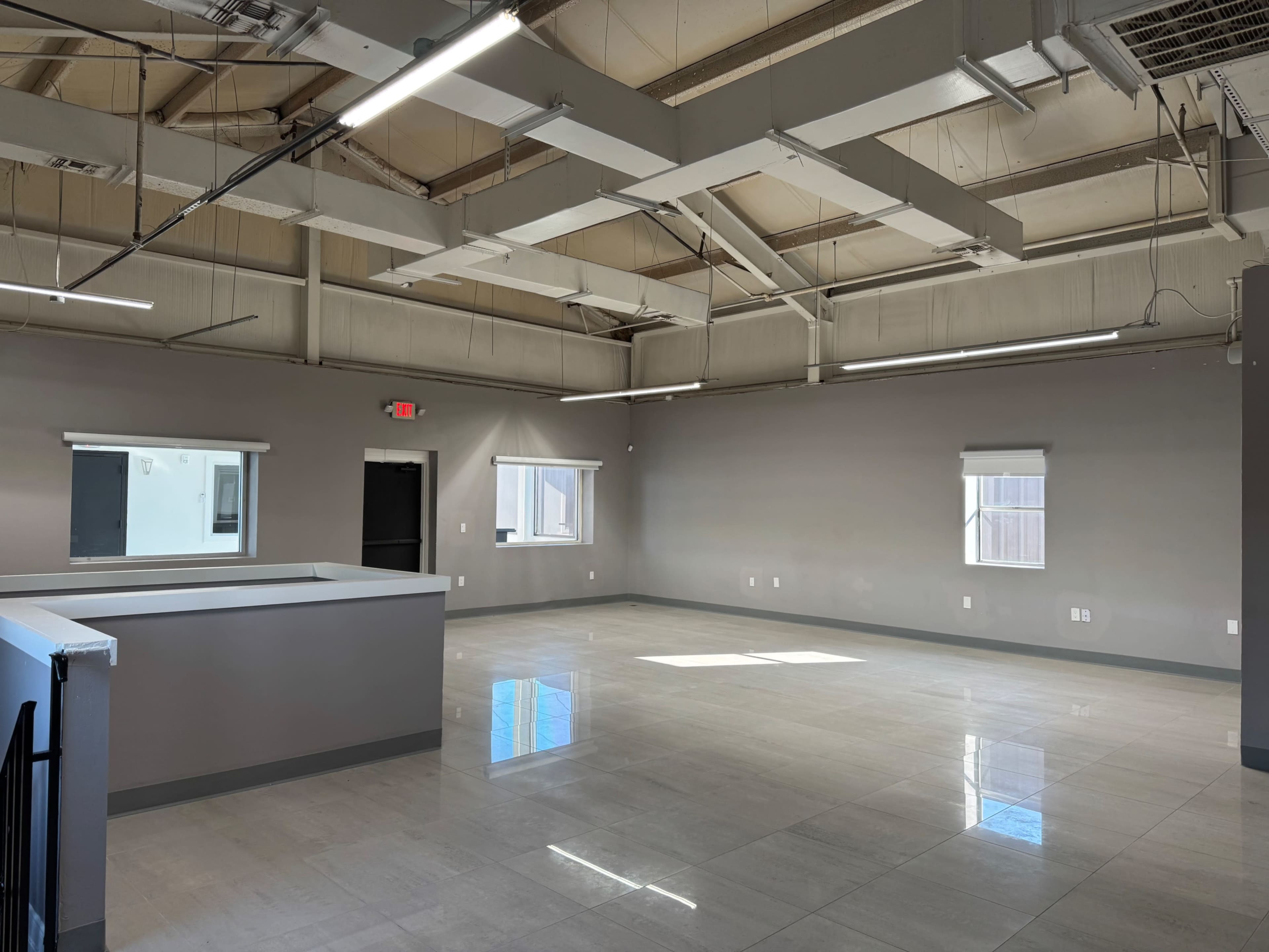 The image shows a spacious, well-lit room with gray walls, tiled flooring, and large windows, featuring a counter in the foreground.