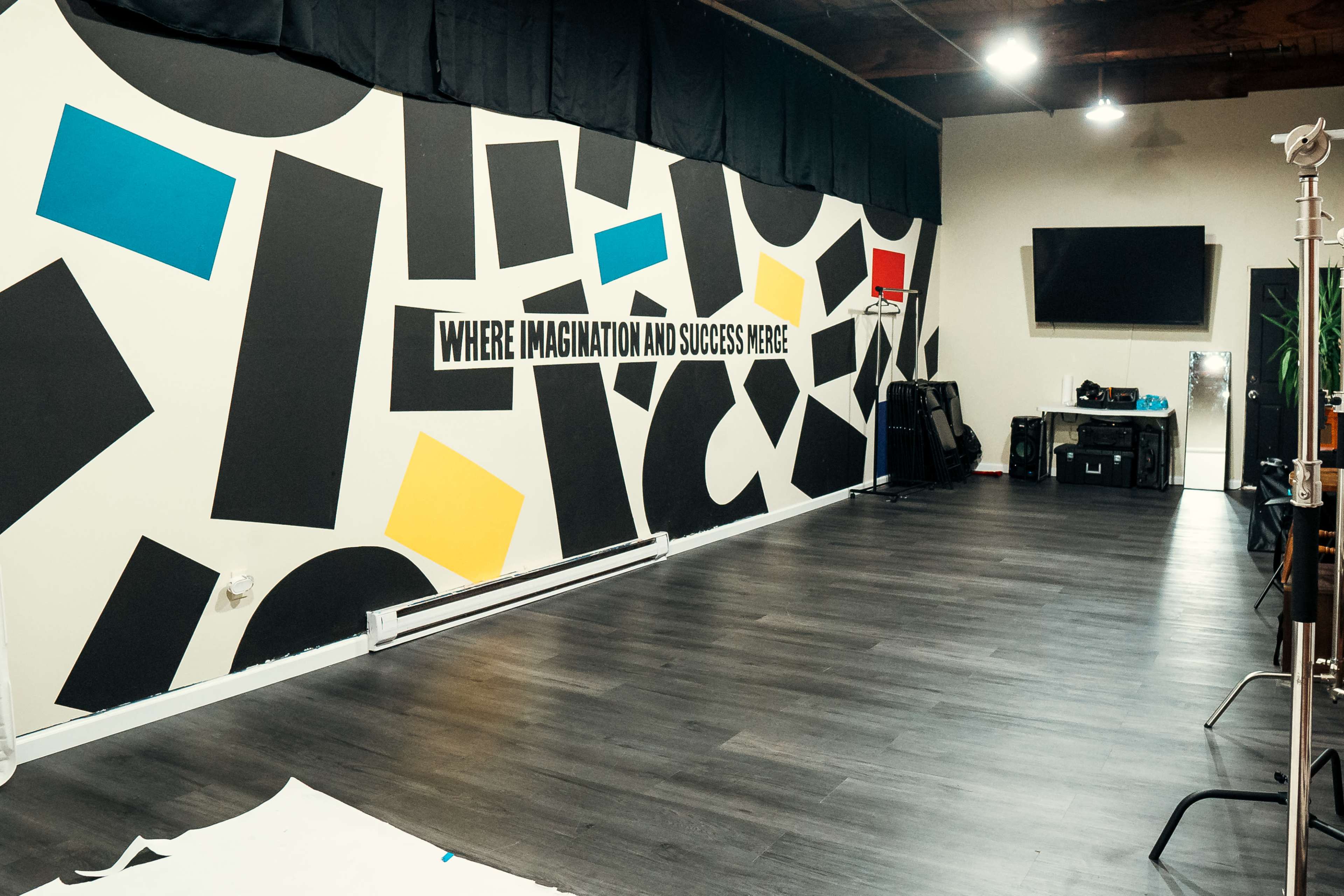 The image shows an empty room with a wall featuring bold black, blue, yellow, and white shapes, along with the phrase "WHERE IMAGINATION AND SUCCESS MEET."