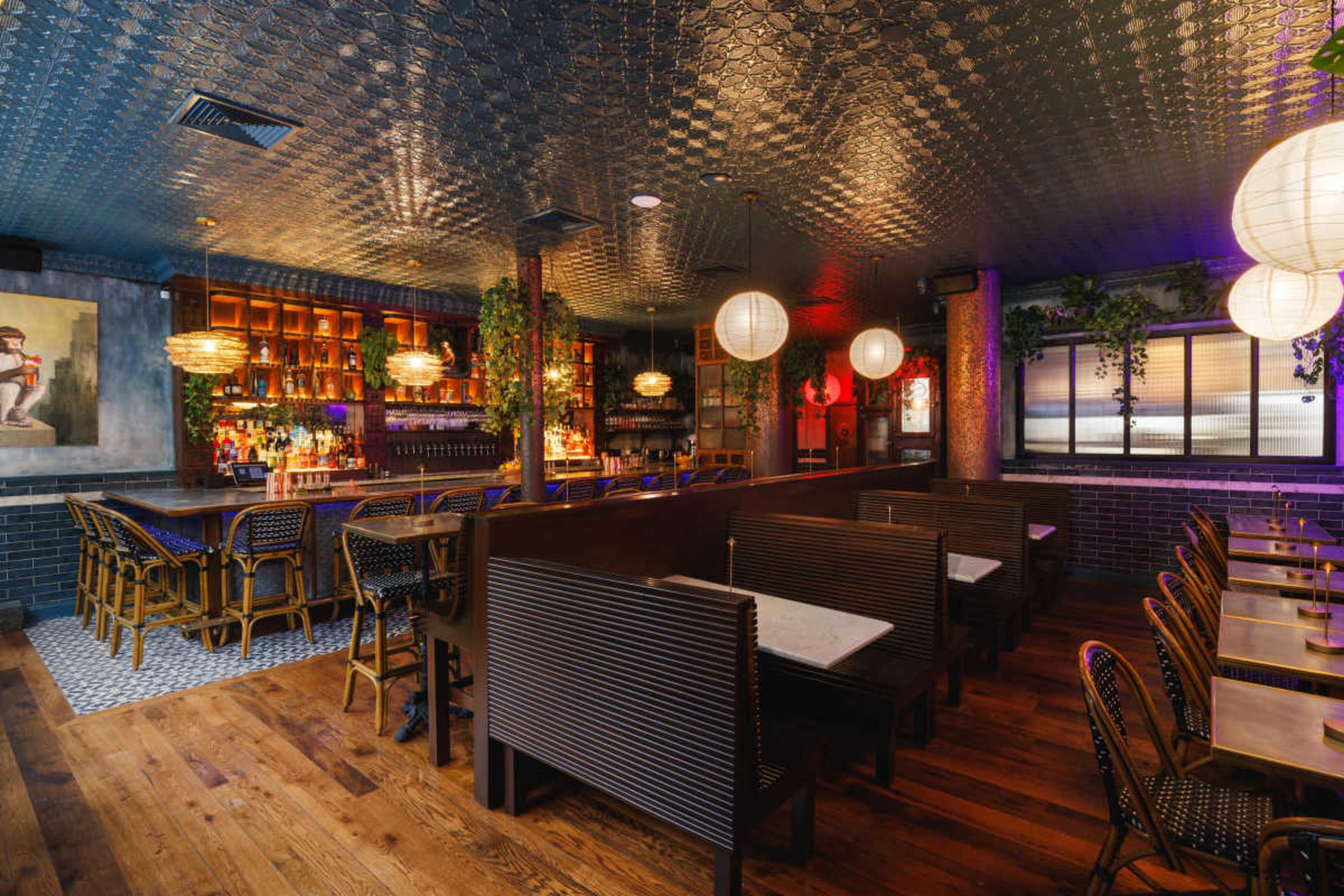 The image shows a dimly lit restaurant interior with wooden flooring, patterned ceiling, and seating booths arranged around a central bar area.