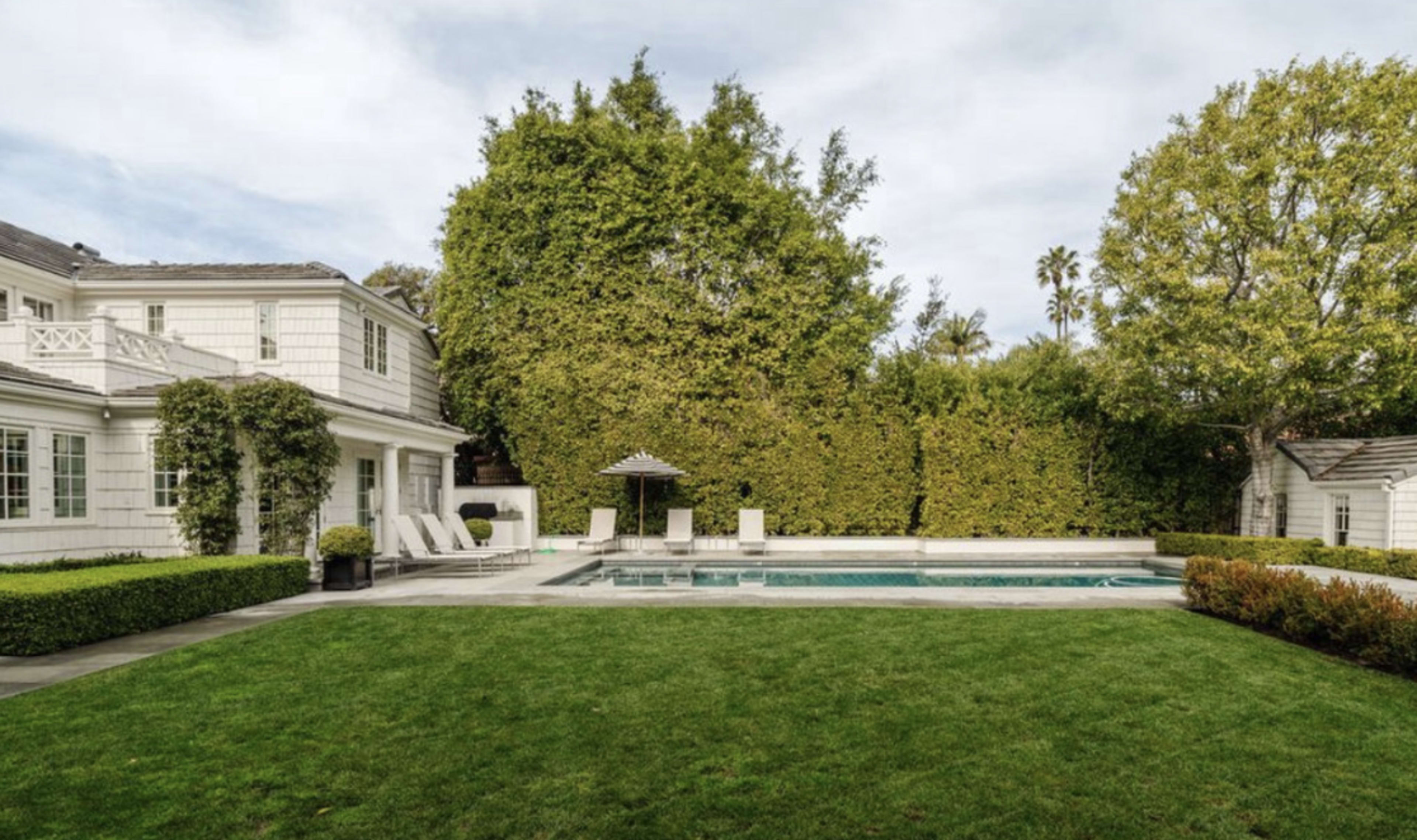 A lush green lawn surrounds a swimming pool, with a house and tall hedges in the background.