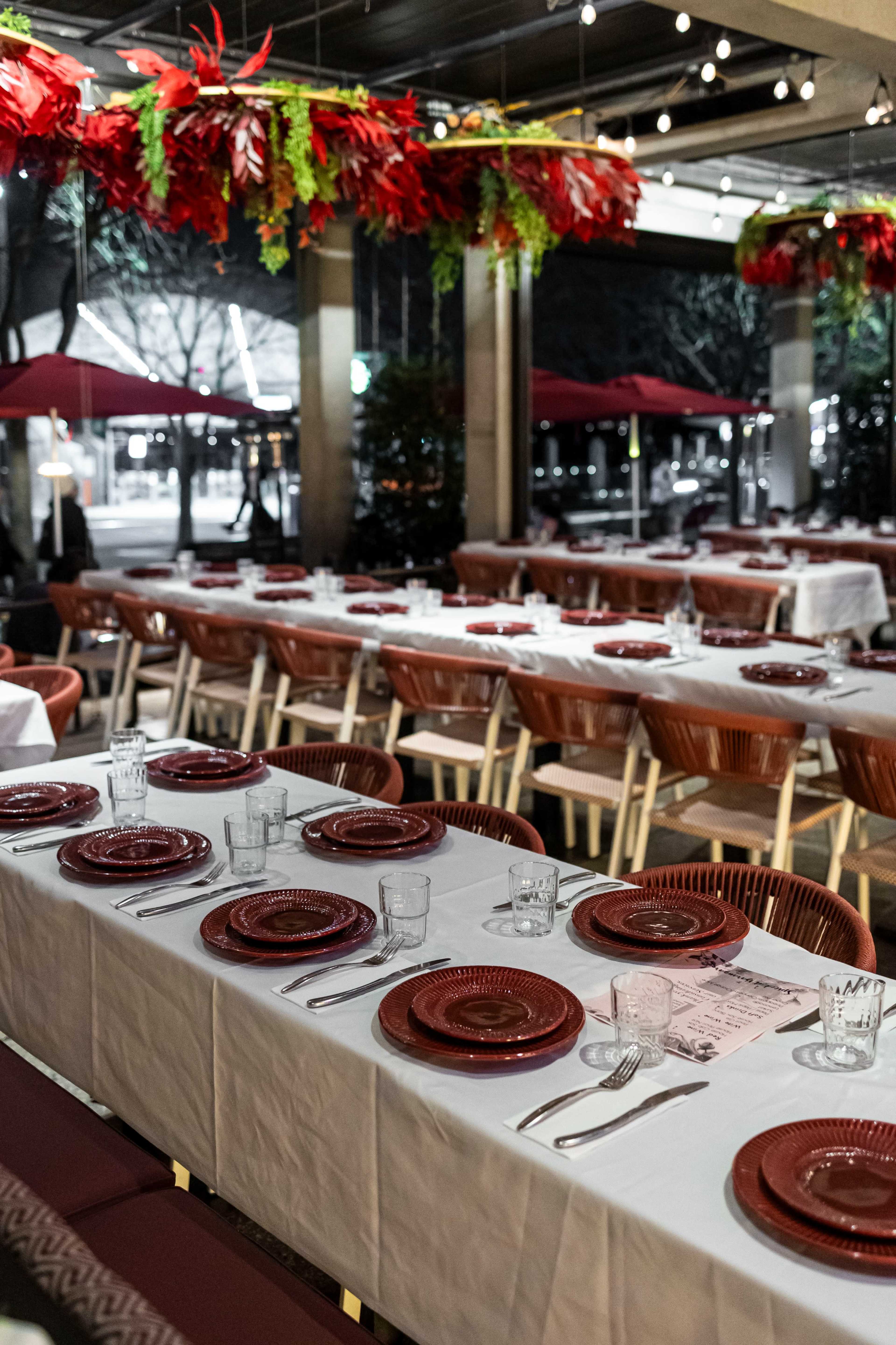 A dining area is set with white tablecloths, red plates, and glassware, featuring decor with hanging greenery and red foliage.