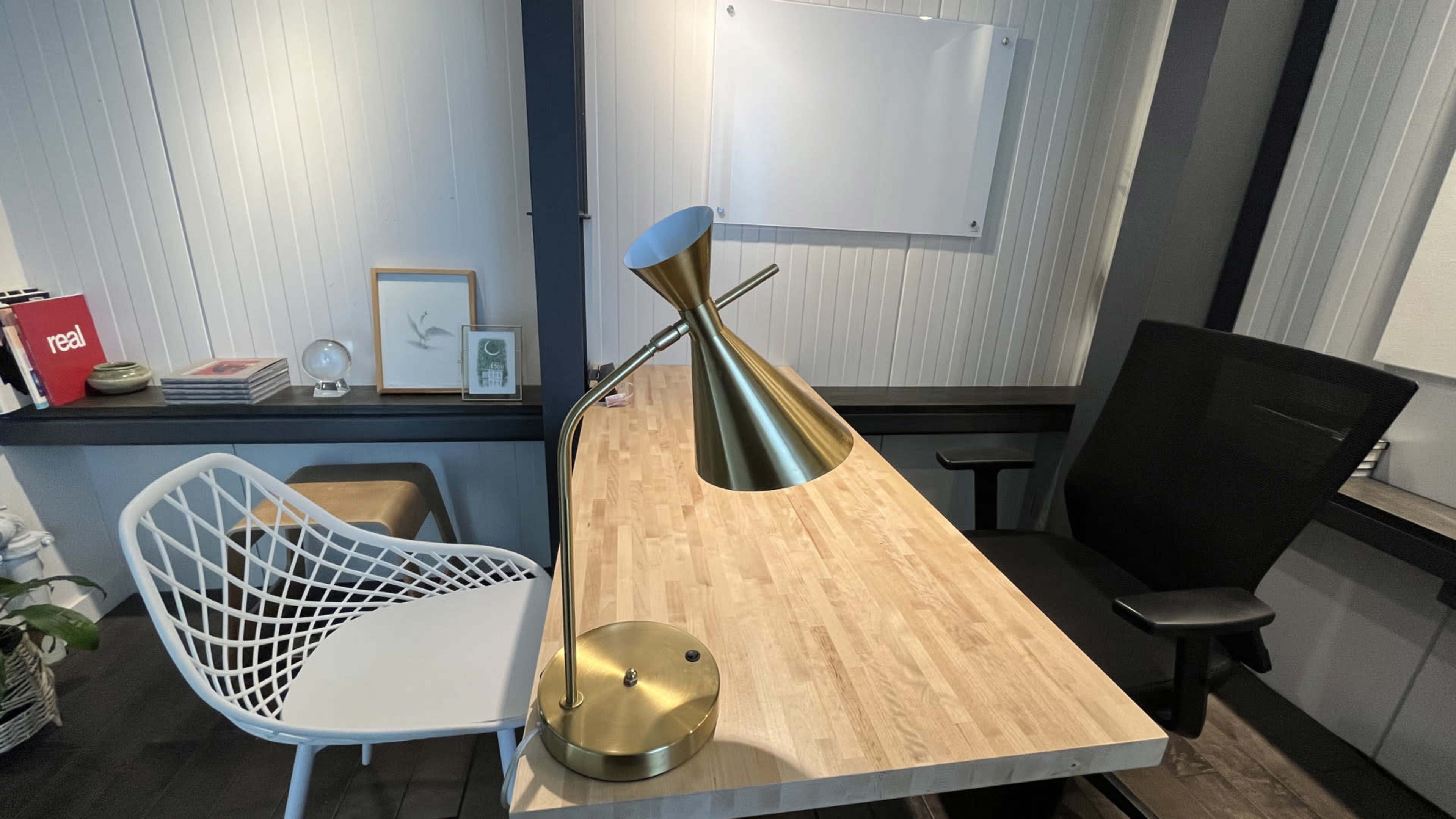 A modern office space features a wooden desk with a brass desk lamp, a white decorative chair, and a black office chair.