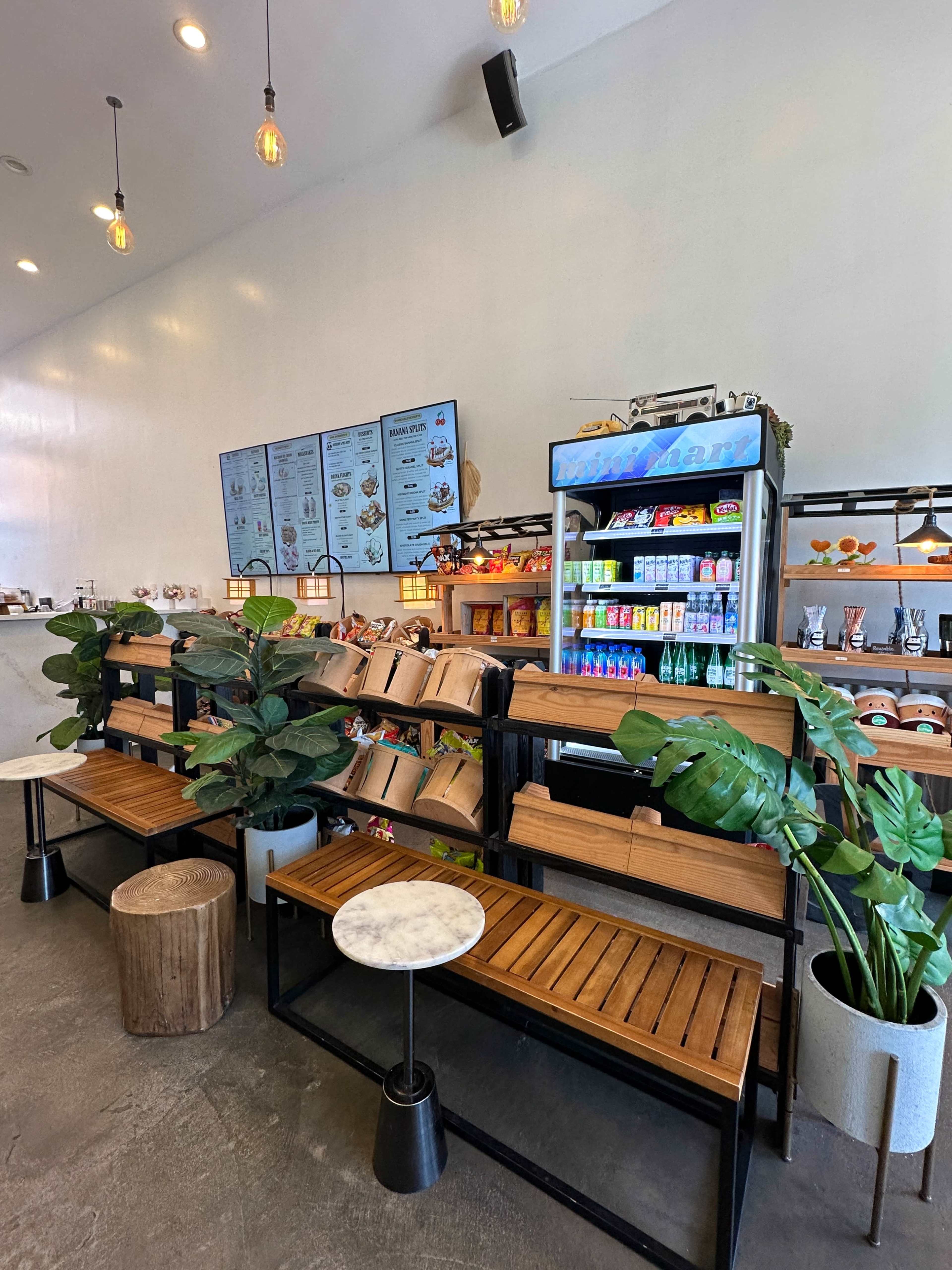 The image shows a modern café interior featuring wooden seating, shelves stocked with snacks and beverages, and decorative plants.