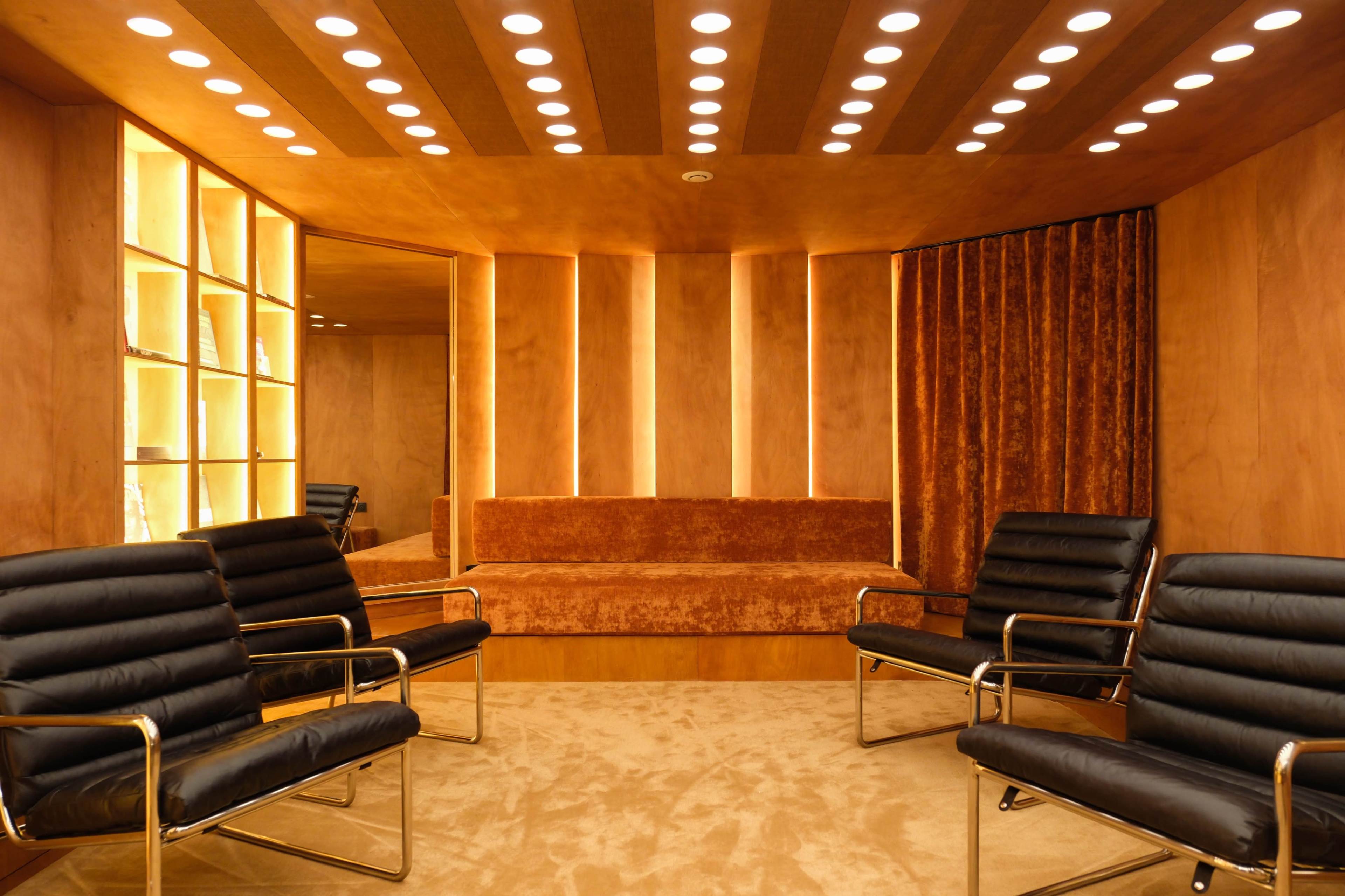 The image shows a modern lounge area with black leather chairs, a plush bench, and warm, illuminated walls.