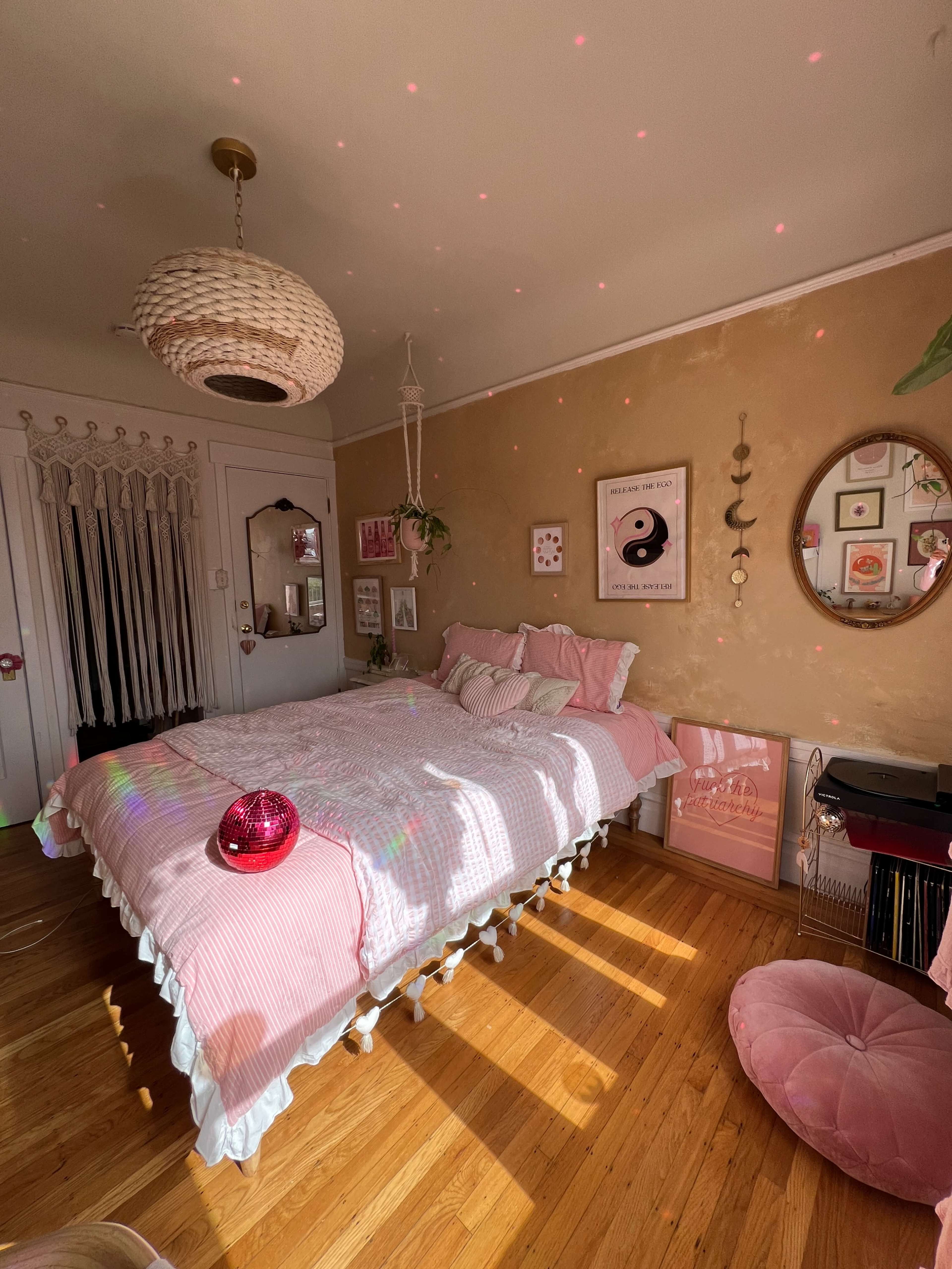 A bedroom features a large bed with pink bedding, a round pink pouf, a hanging light fixture, and wall art, illuminated by sunlight streaming through the windows.