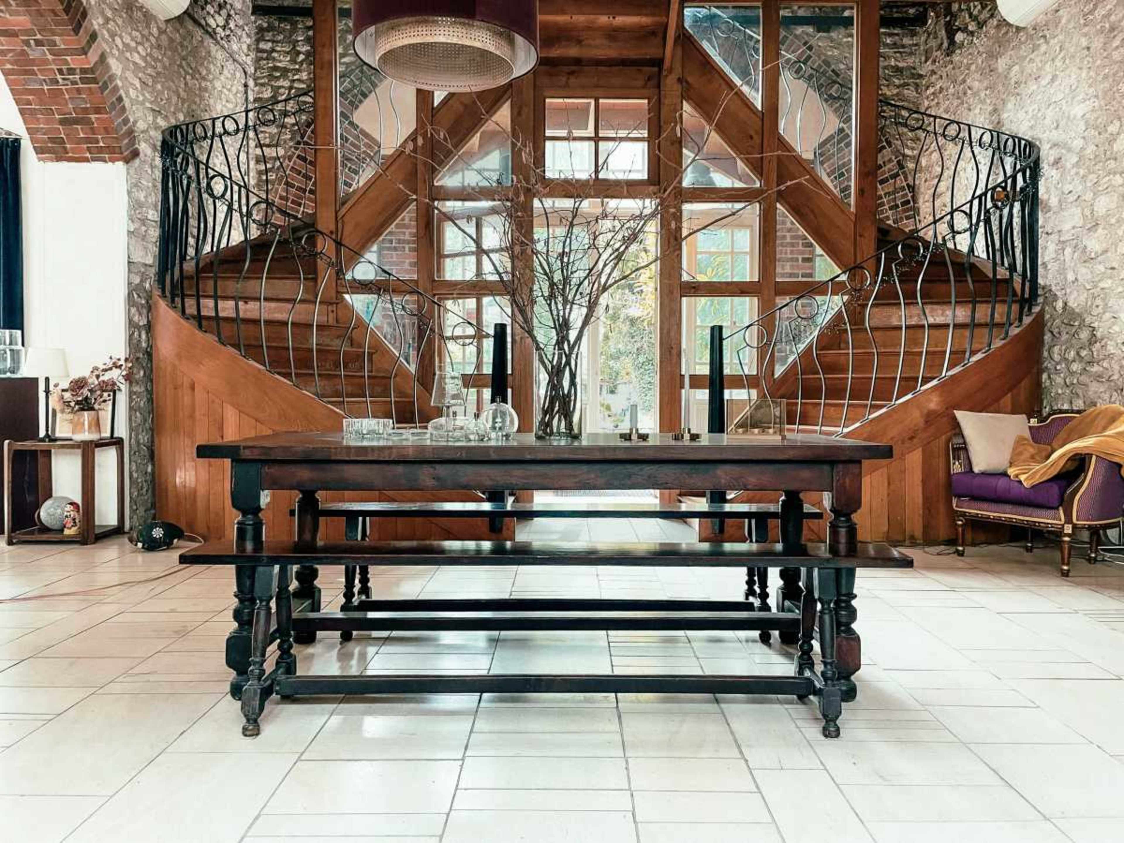 A wooden table is placed in the center of a spacious entryway featuring a grand staircase with decorative railings.