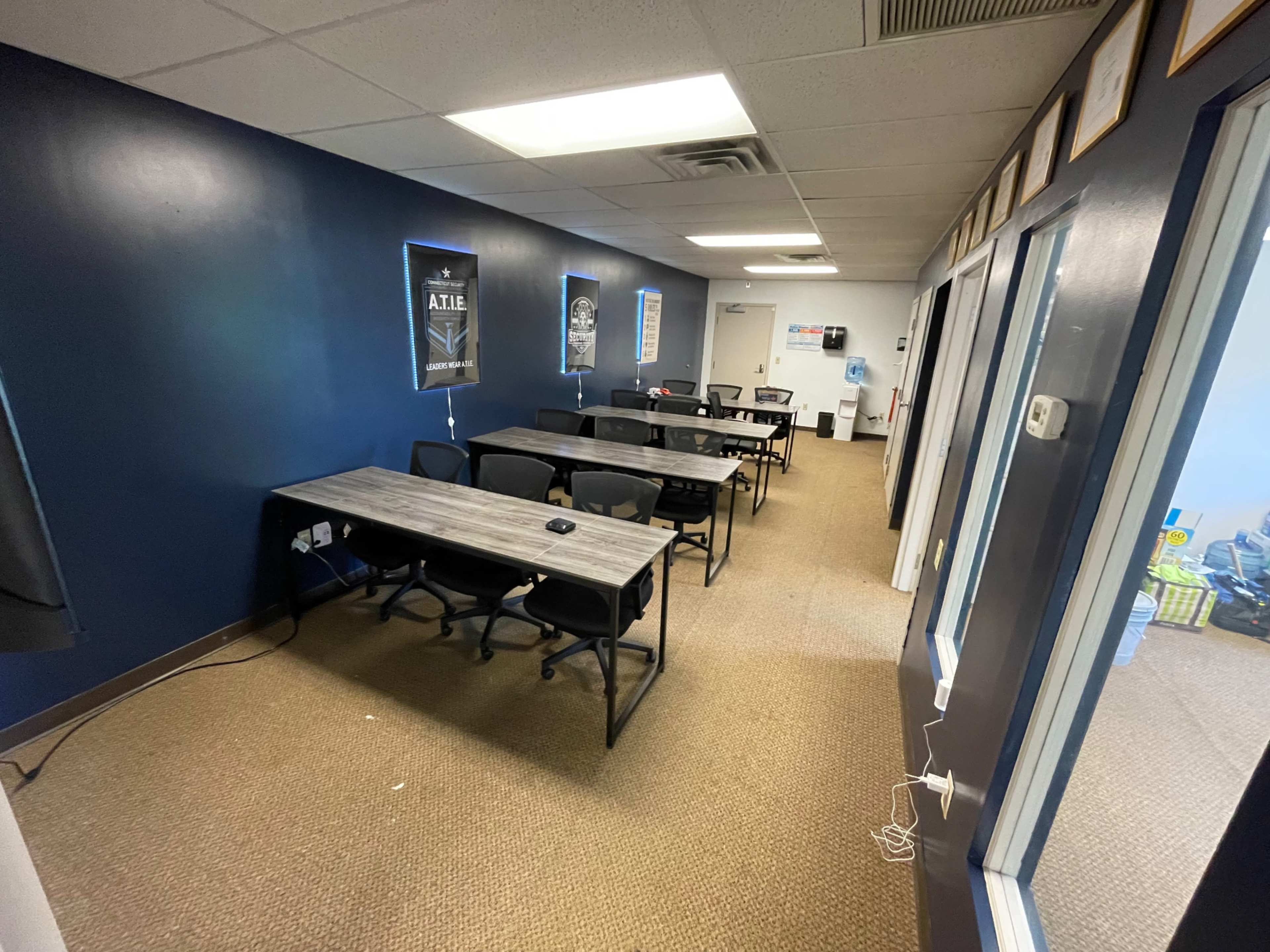 The image shows a long, narrow room with several gray tables and black chairs arranged in rows along a dark blue wall.