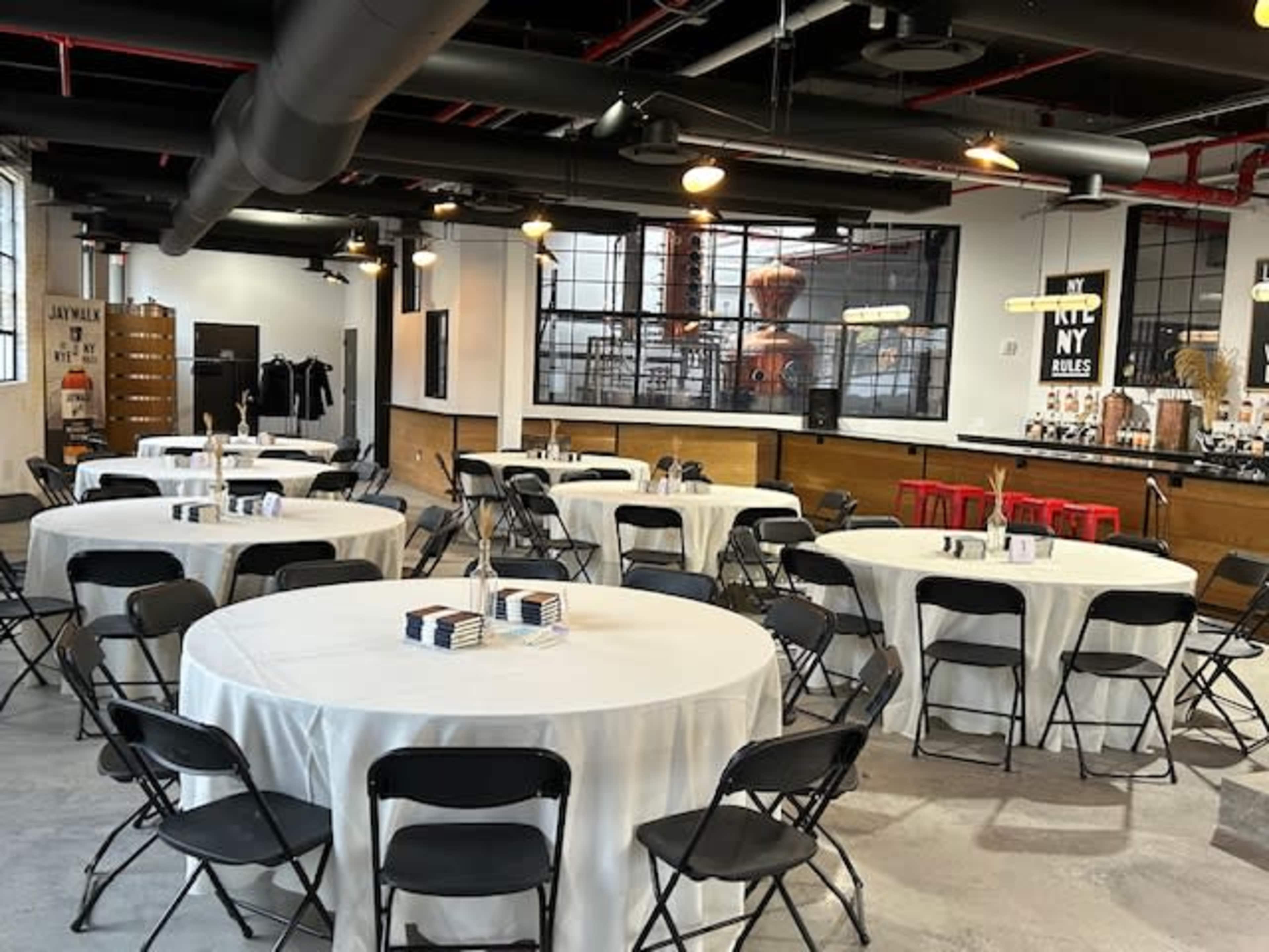 A neatly arranged event space with several round tables covered with white tablecloths and black chairs, featuring a bar area in the background.