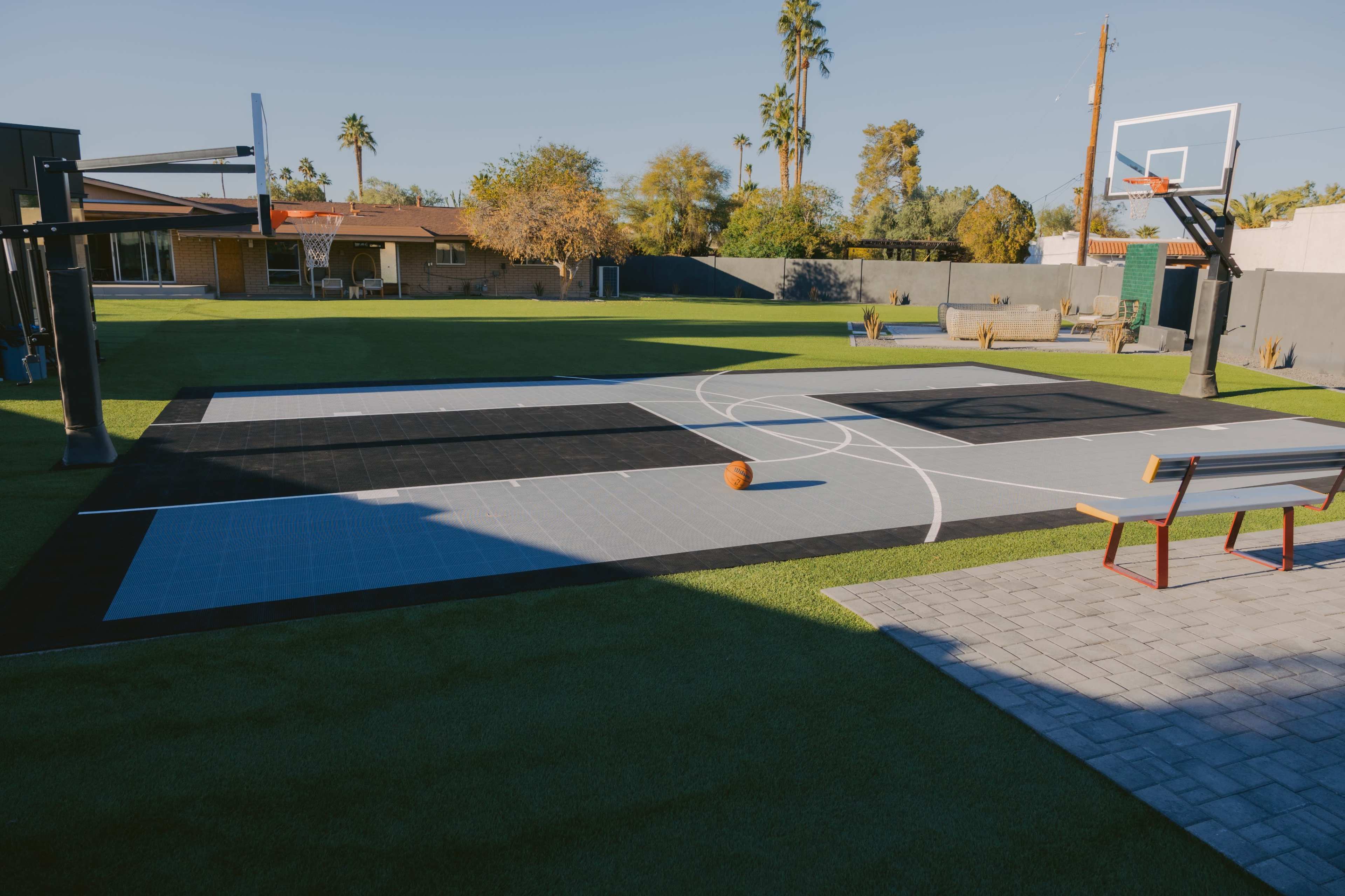 The image shows a basketball court with a multi-colored surface, set in a spacious backyard with palm trees and a residential building in the background.