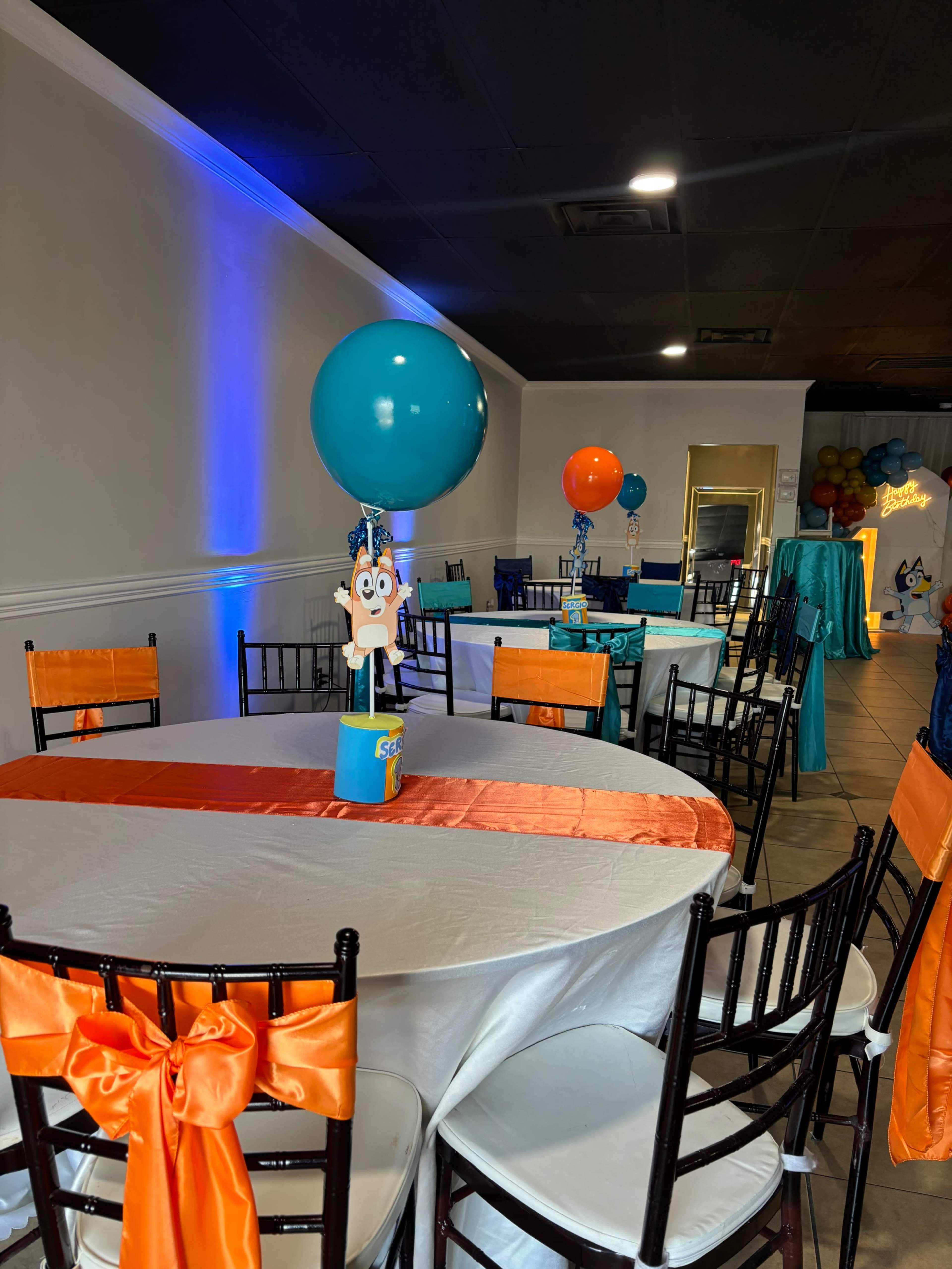 The image shows a decorated event space with tables set for a celebration, featuring colorful balloons and themed centerpieces.