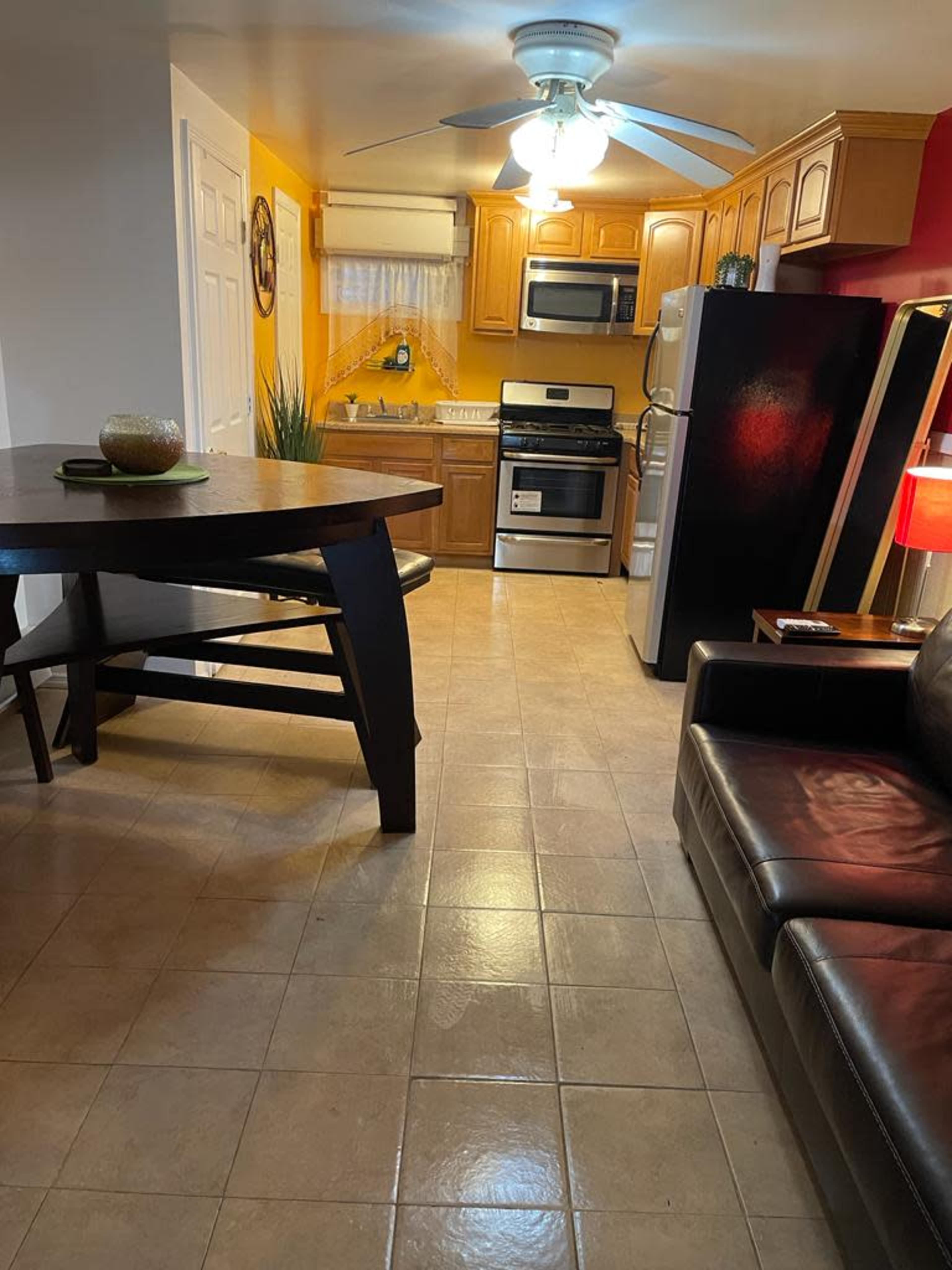 A small kitchen area with wooden cabinets, a stove, a refrigerator, and a table set beside a couch in a brightly lit room.